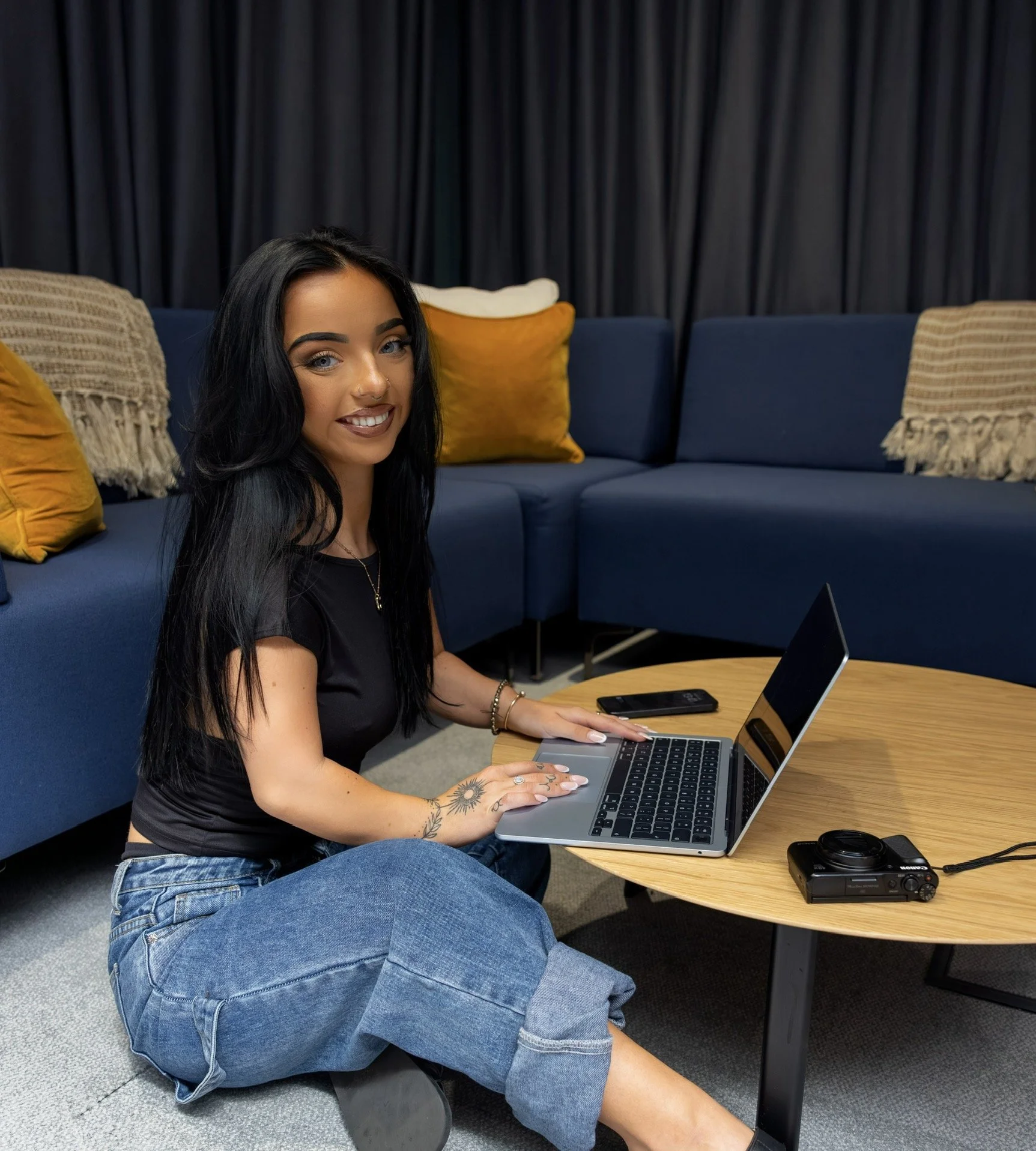 Young woman with long black hair, sitting on the floor, working on a laptop at a wooden coffee table in a cozy living room with blue couches and yellow pillows.