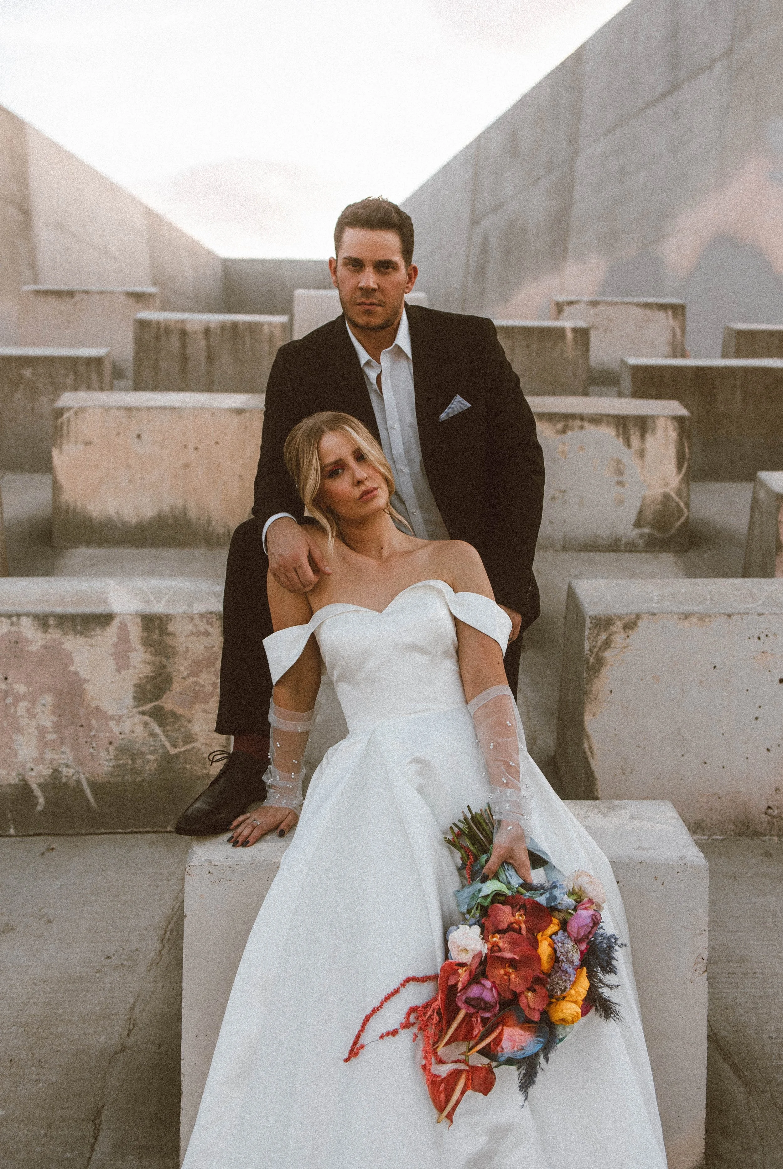 A man in a black suit and a woman in a white wedding dress sitting on concrete blocks. The woman holds a colorful bouquet, and the man has his hand on her shoulder, both posing for a wedding photo in an outdoor, industrial-looking setting with concrete structures.