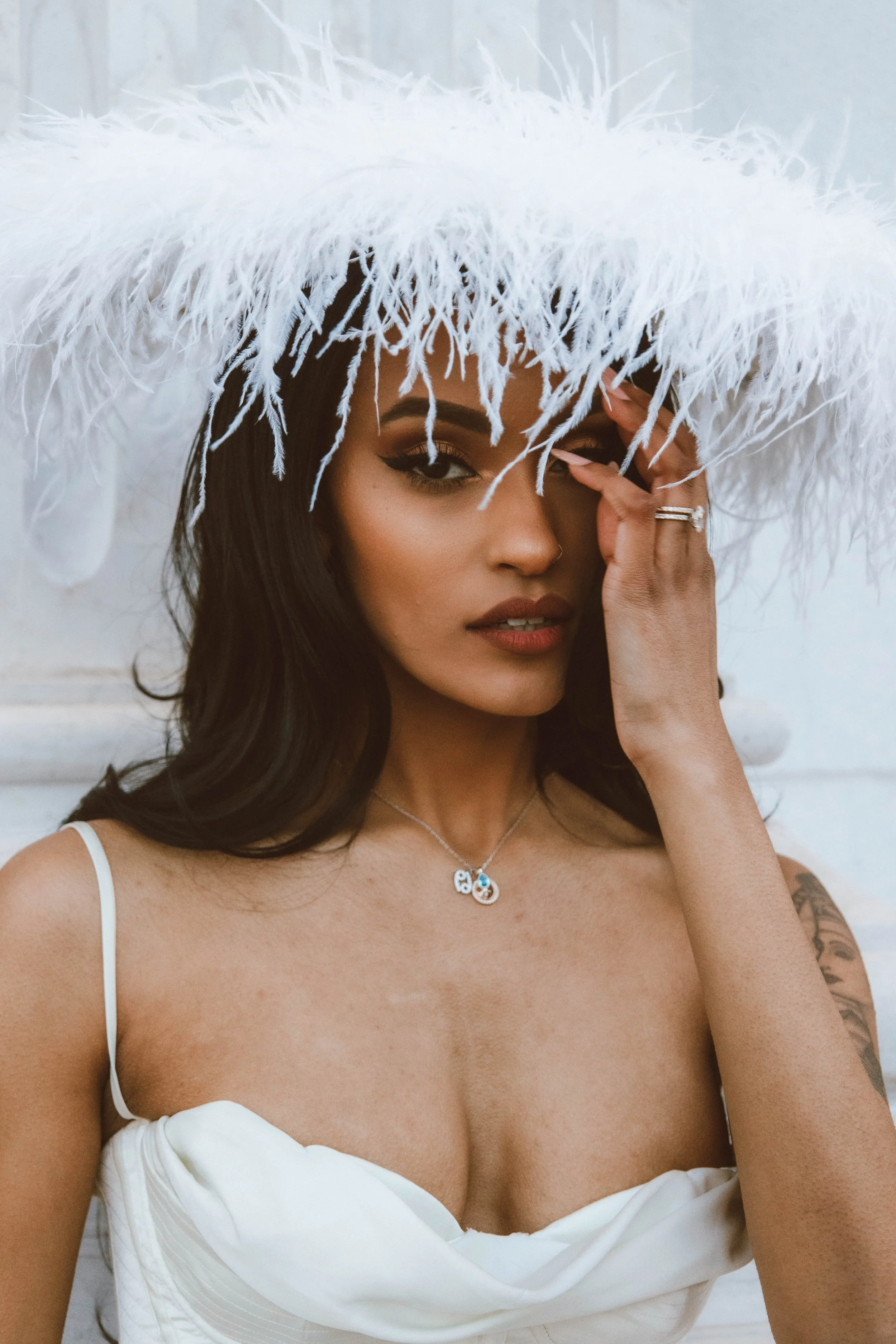 A woman wearing a large white feathered hat and white dress, with necklace, rings, and tattoo, looking at the camera.