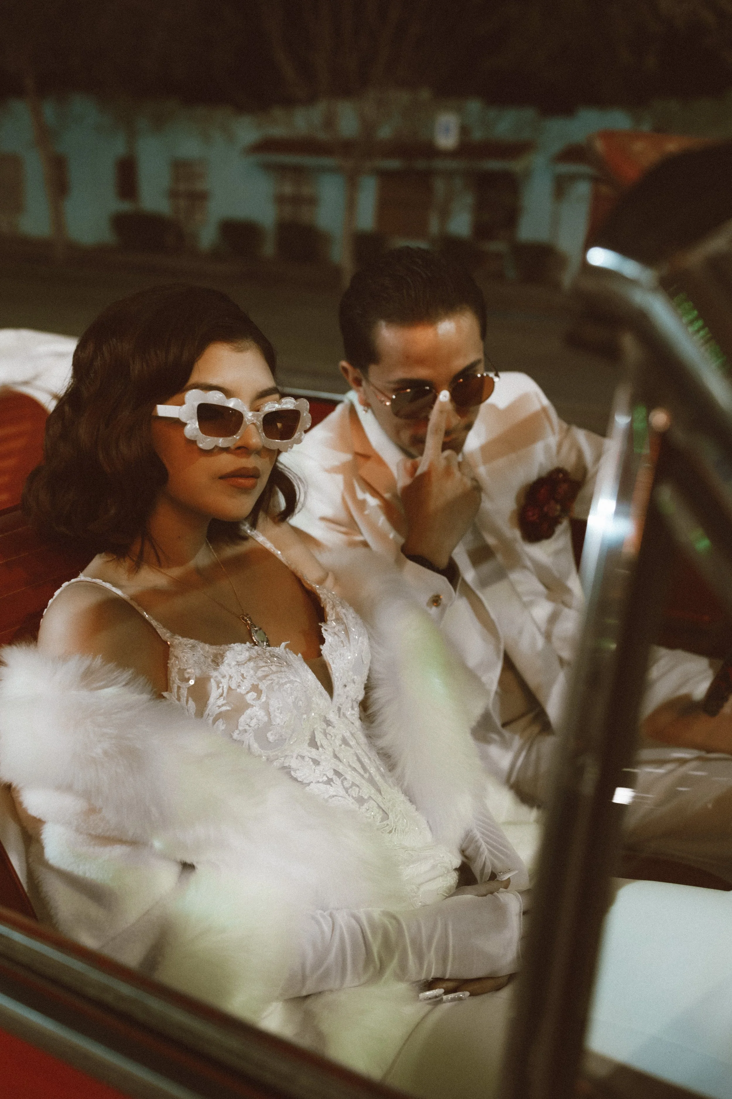 A woman in a white dress and white gloves sitting in a vintage red car, wearing white sunglasses with a decorative frame, with a man in a white suit with sunglasses, holding his finger near his nose, sitting next to her.