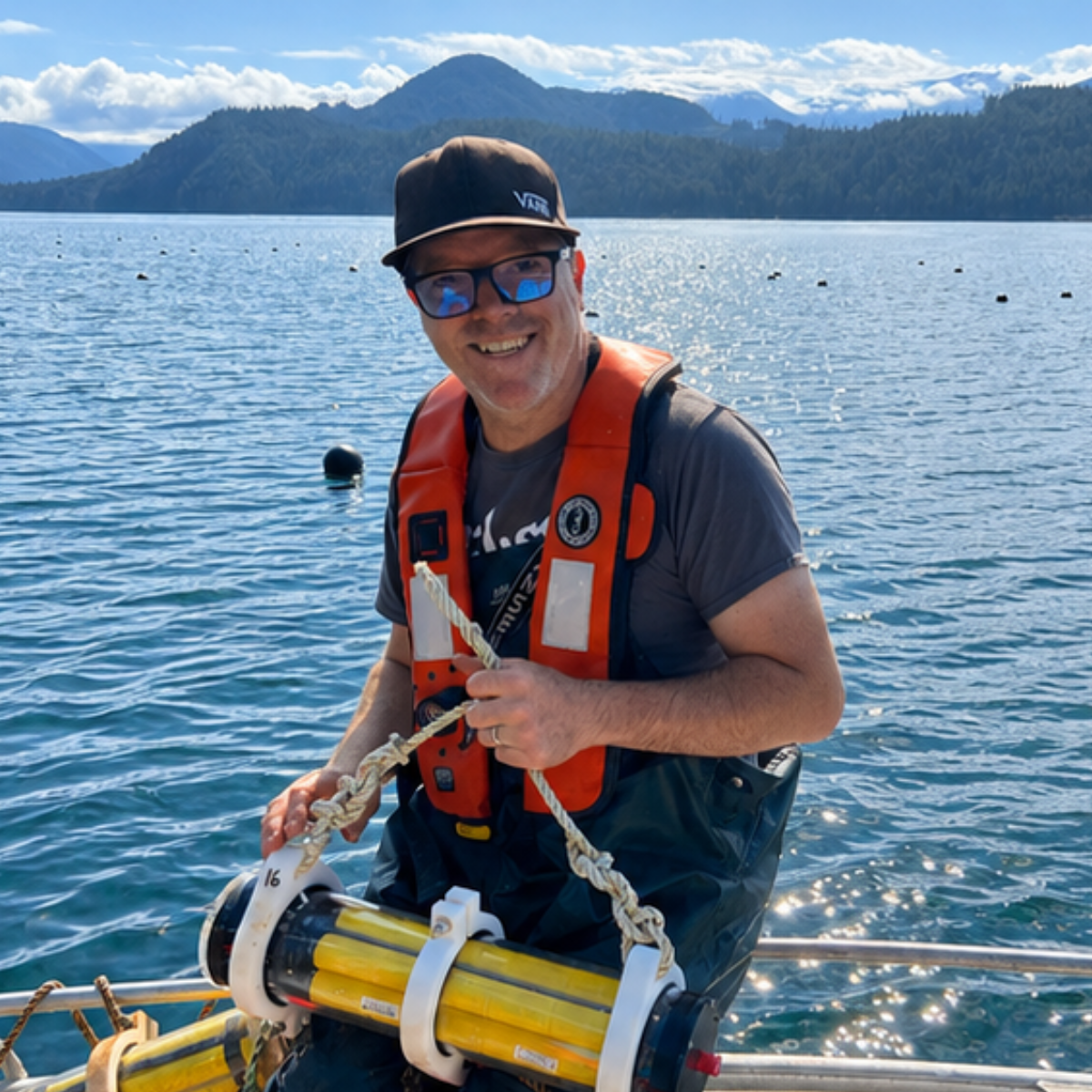 A man in sunglasses and a black cap smiling on a boat on a lake, holding a piece of scientific equipment with yellow tubes, with mountains and blue sky in the background.