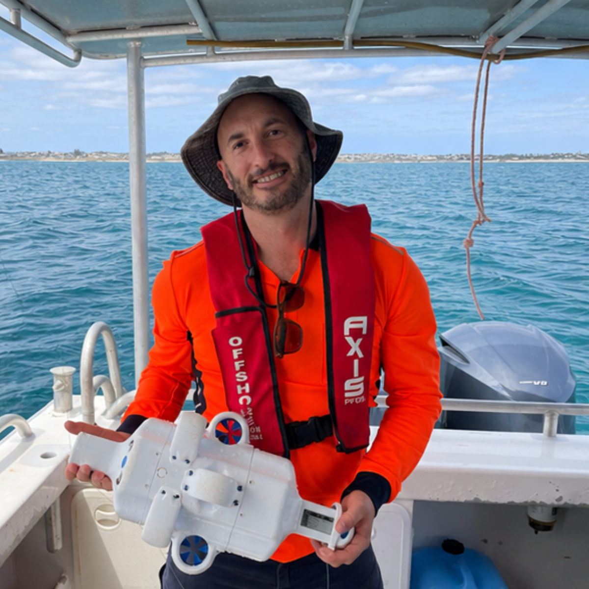 A man in a red life jacket and orange shirt on a boat holding a scientific instrument, with ocean and coastline in the background.