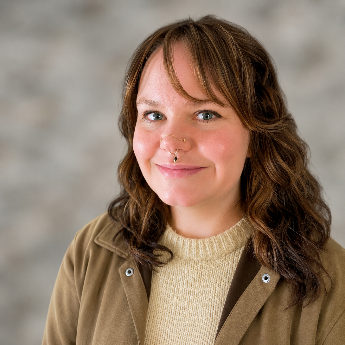 A woman with wavy brown hair and blue eyes smiling, wearing a beige sweater and a tan jacket, with facial piercings including a nose ring and a septum ring, against a blurred gray background.