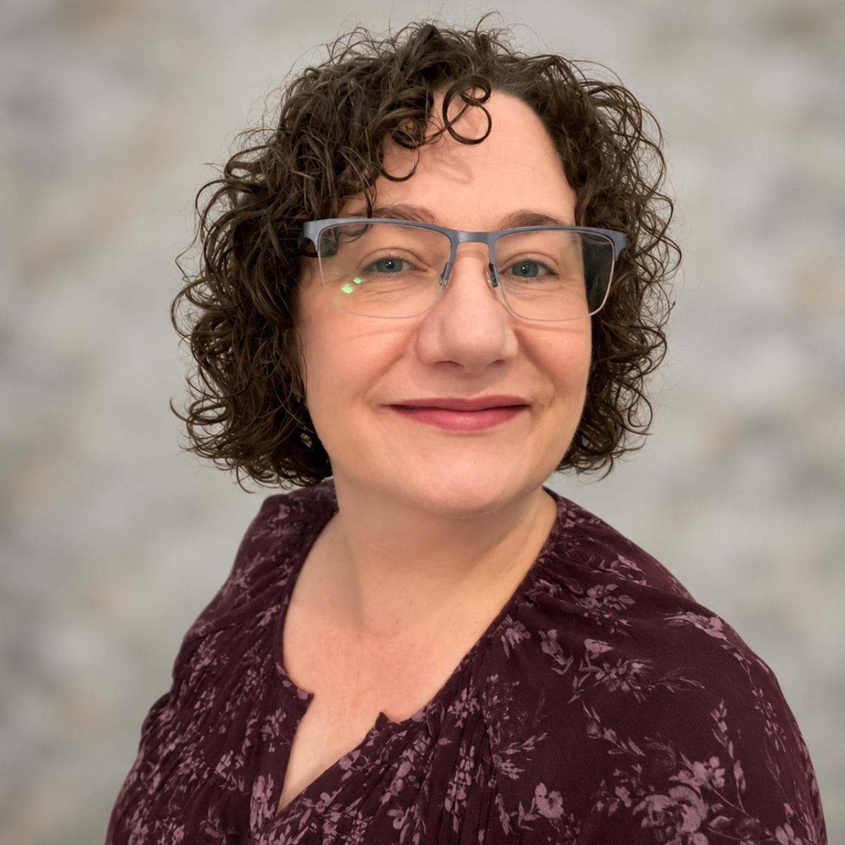 A woman with short, curly brown hair and glasses, smiling at the camera, wearing a burgundy floral blouse.