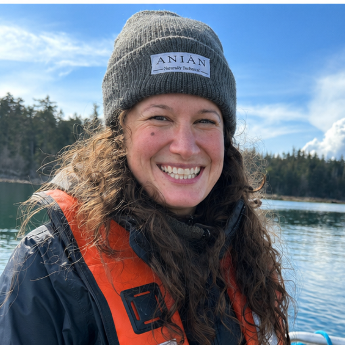 A smiling woman wearing a gray beanie with a patch, an orange life jacket, and a jacket, standing outdoors near water with a forest in the background on a partly cloudy day.