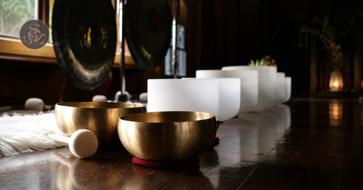 A row of singing bowls, with two brass bowls in the front and three white bowls behind, on a wooden floor near a window with natural light in a cozy indoor setting.