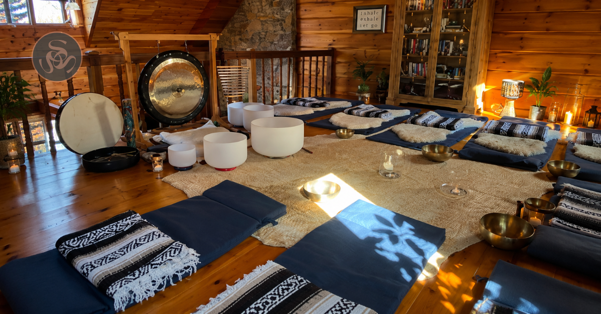 A cozy meditation or sound healing room with floor cushions, singing bowls, mallet, a gong, and candles arranged on a wooden floor and walls.