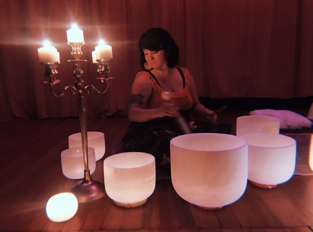 A woman playing a black metal handpan drum in a dimly lit room illuminated by candles and soft lights, sitting on the floor surrounded by white lit-up bowls.