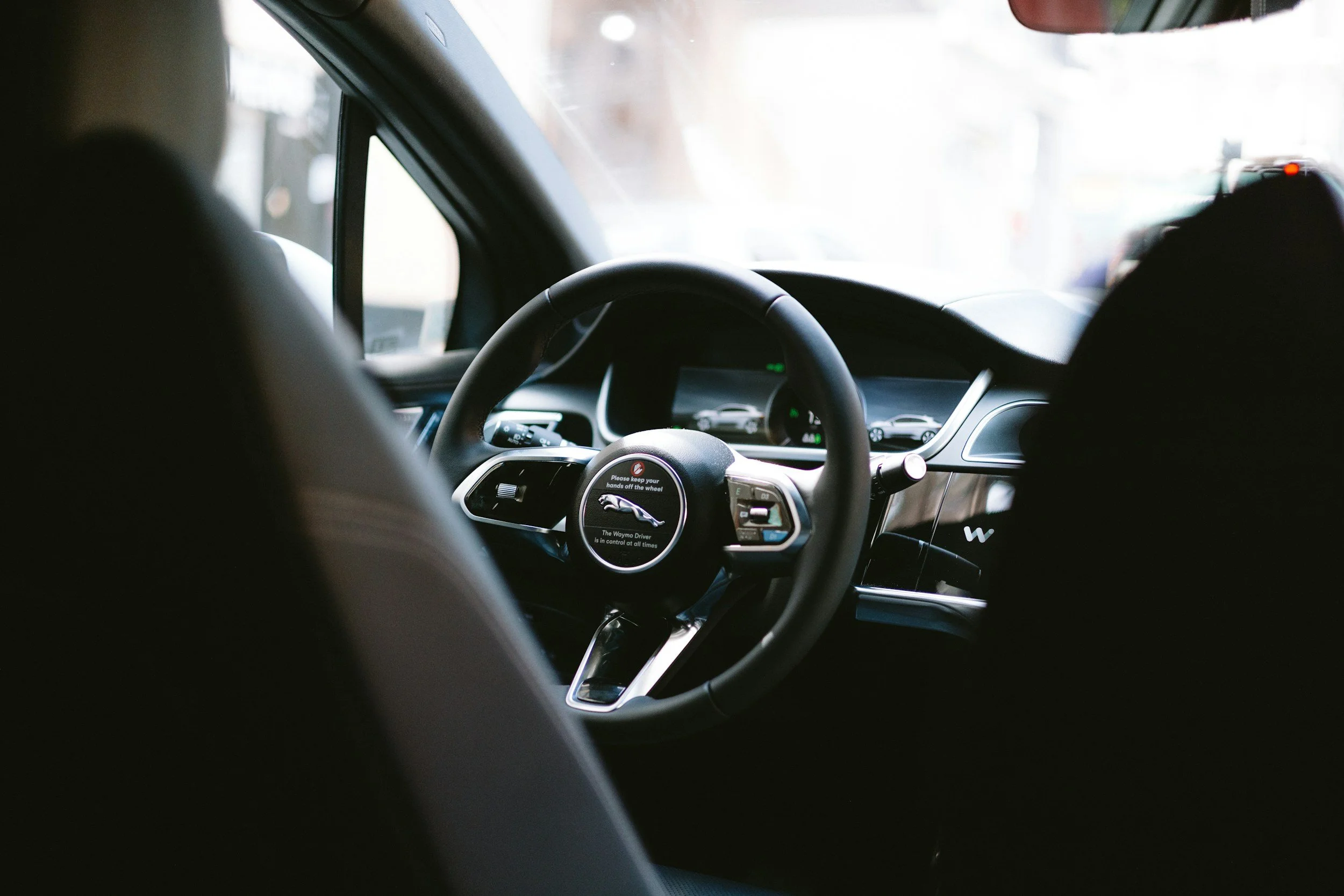 The interior of a Tesla vehicle from the back seat perspective, showing the steering wheel with Tesla logo, digital dashboard, and windshield with bright outdoor scene.