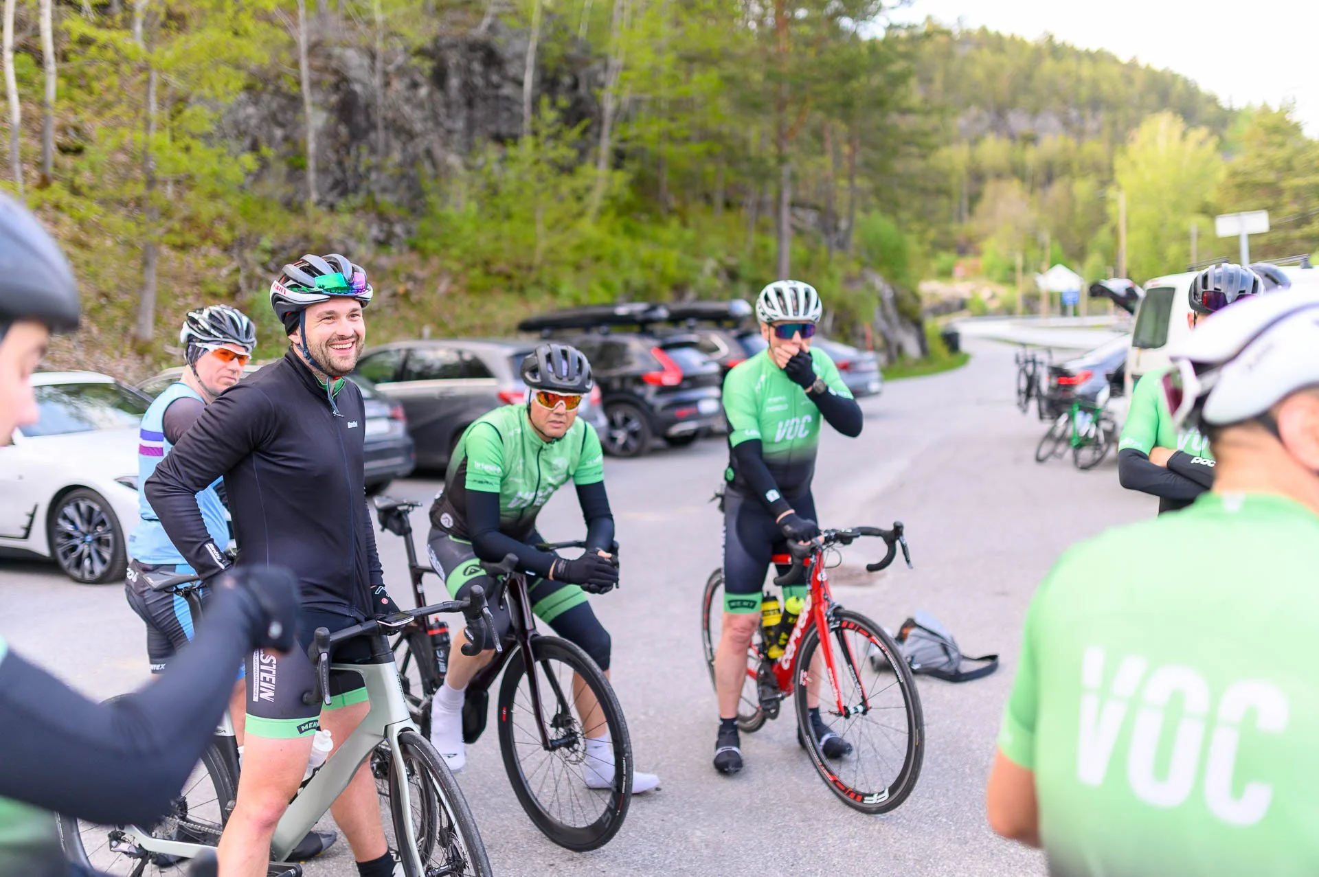 En gruppe syklister iført hjelm og sykkelklær samles på et parkeringsområde, omgitt av biler og grønne trær i bakgrunnen.
