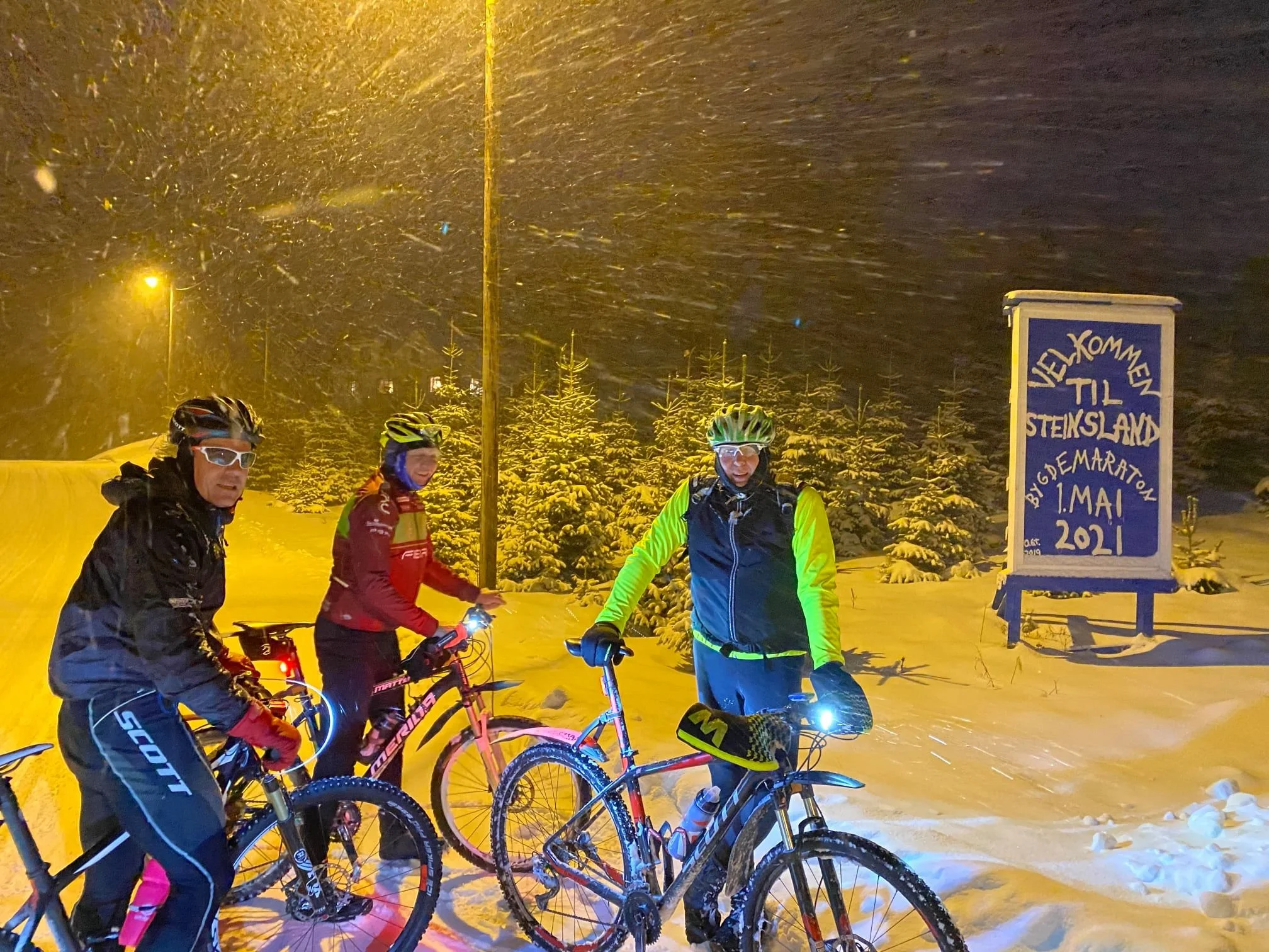 Tre syklister i vinterlandskap om natten med snøvær, foran en skilt som ønsker velkommen til Steinsland. En gruppe syklister er utstyrt med hjelmer og refleksutstyr.