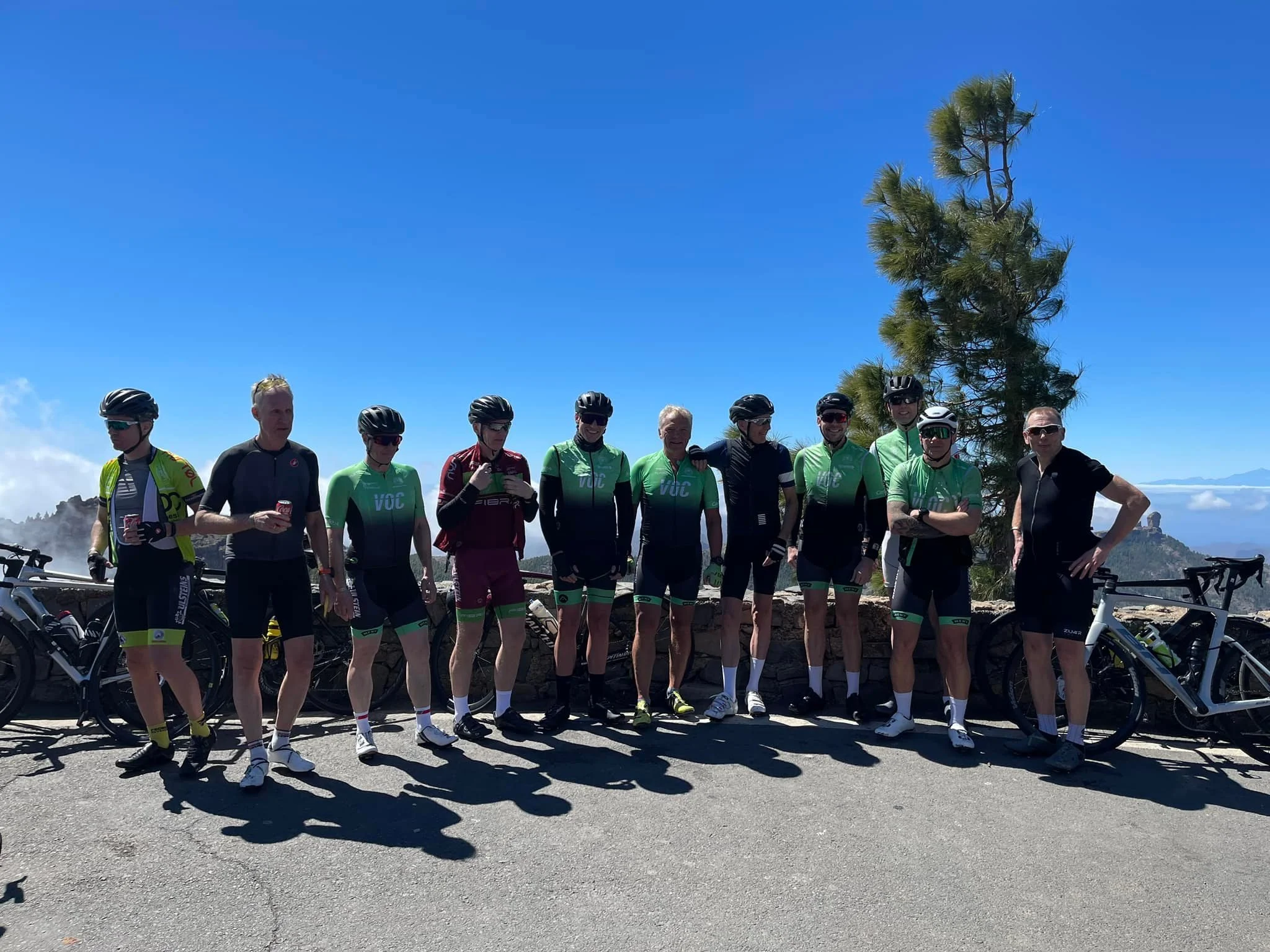 En gruppe syklister står sammen på en vei med fjell og blå himmel i bakgrunnen, noen holder drikke, og flere har på seg sykkelhjelmer og sykledrakter.