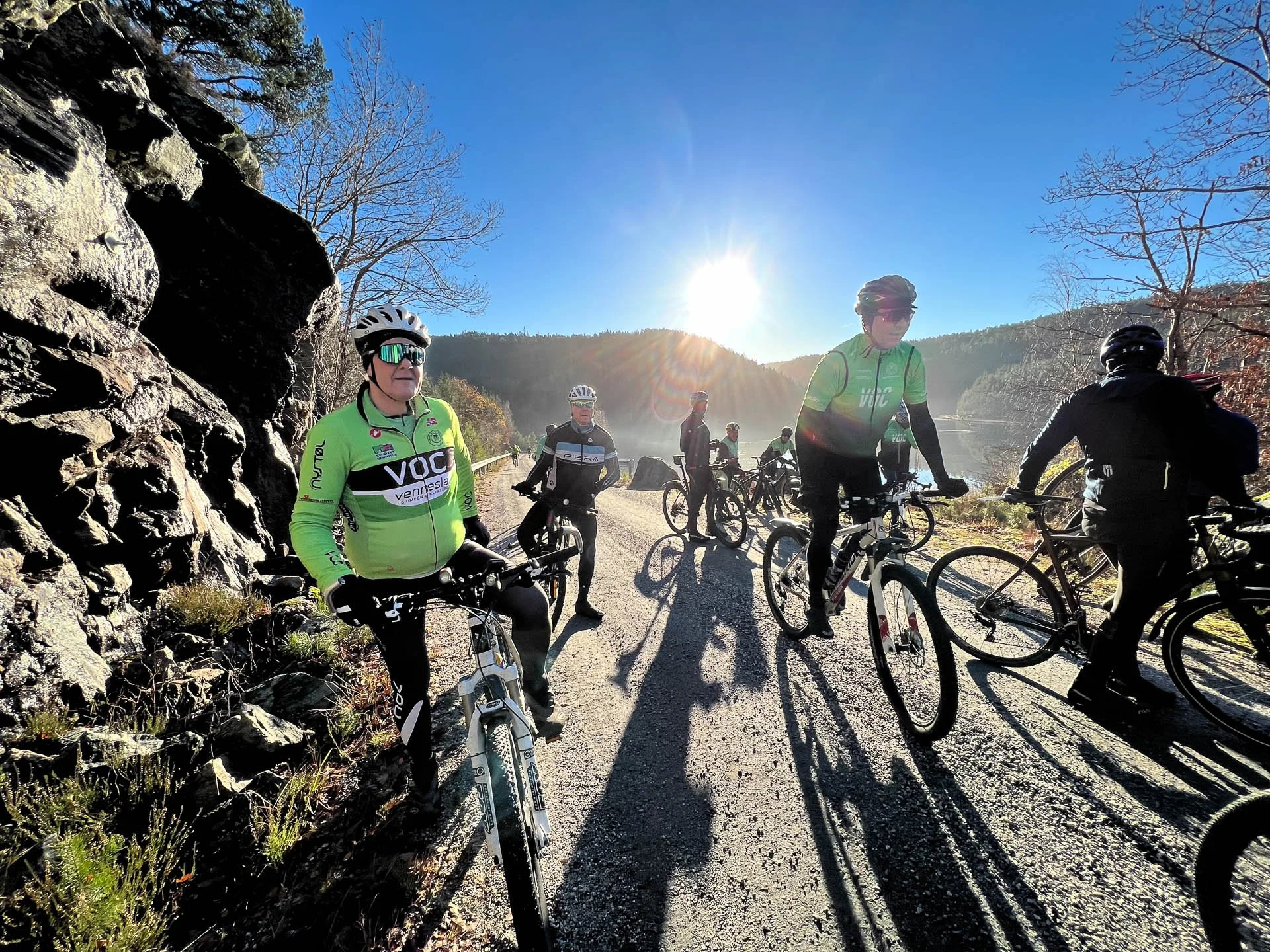 Syv syklister i solskinn på en fjellvei med tre på hver side, fjell og skog i bakgrunnen og klar blå himmel.