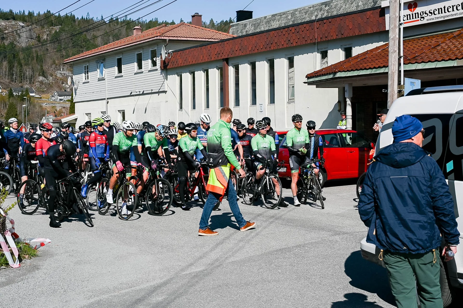 Syklister som venter ved startstreken i en liten norsk by med hus i bakgrunnen og noen biler.