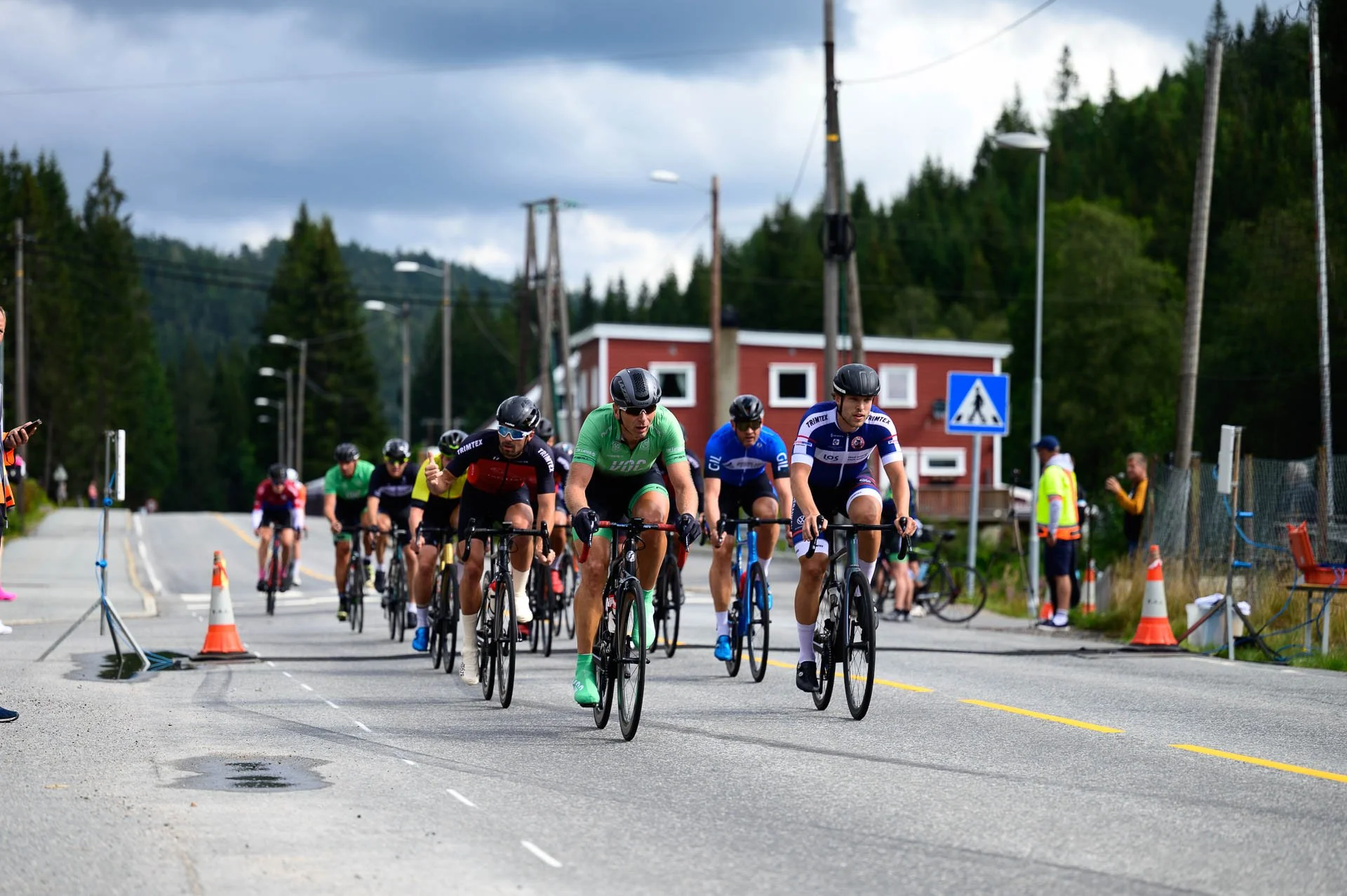 Syklister i et sykkelritt på landevei med hus, treer og veiskilt i bakgrunnen, under en overskyet himmel.