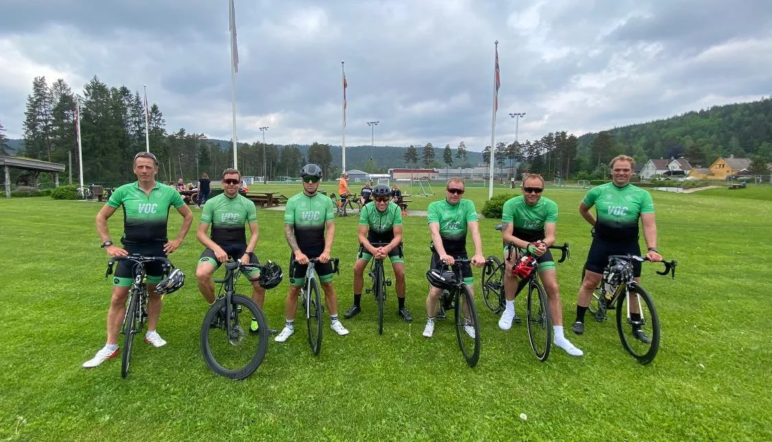 Syv syklister i grøn klubbtrøye står med sykler på en gressbane med flagg og fotballfelt i bakgrunnen.