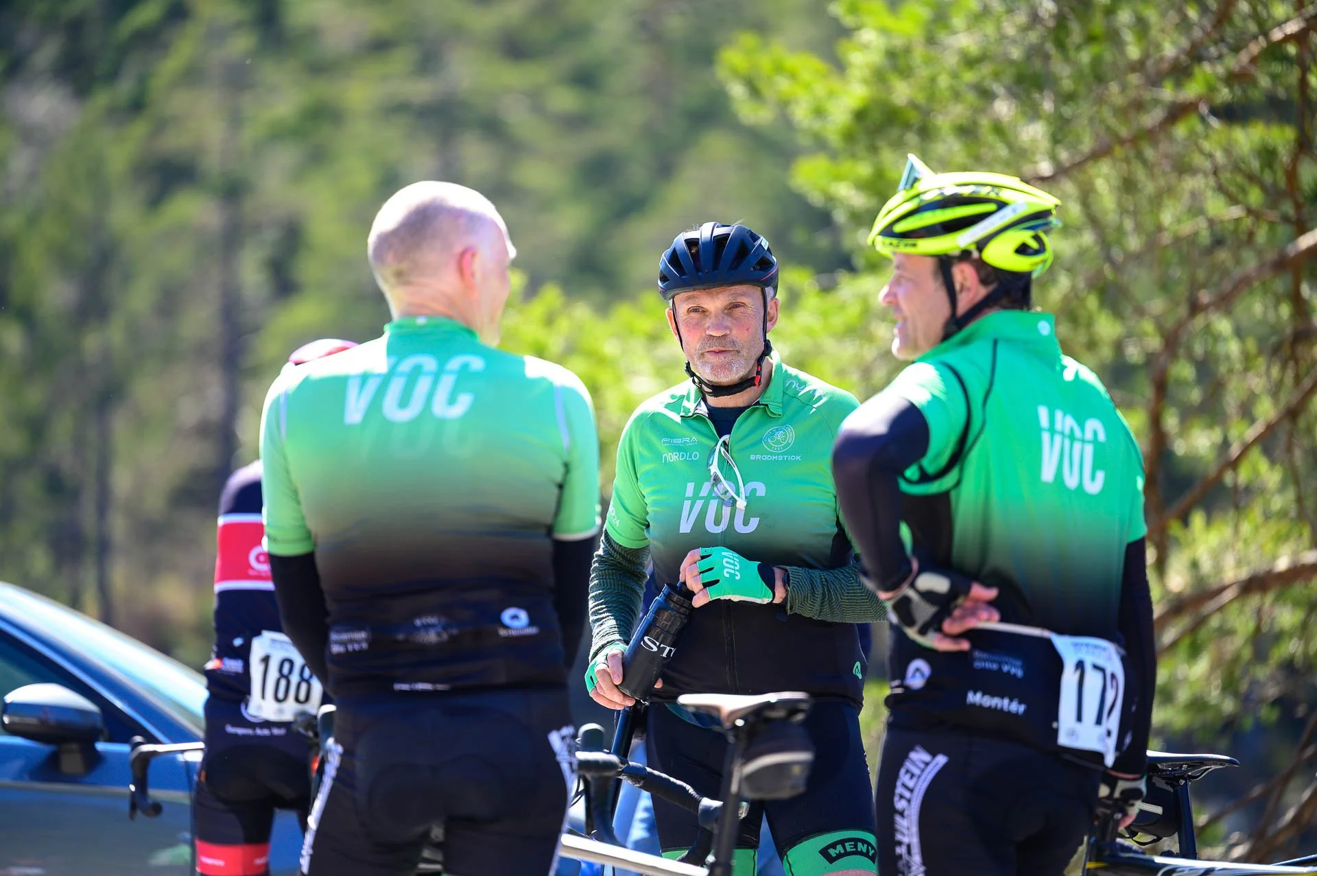 Tre personer i sykkelutstyr snakker sammen utendørs, med en bil og grønn vegetasjon i bakgrunnen.