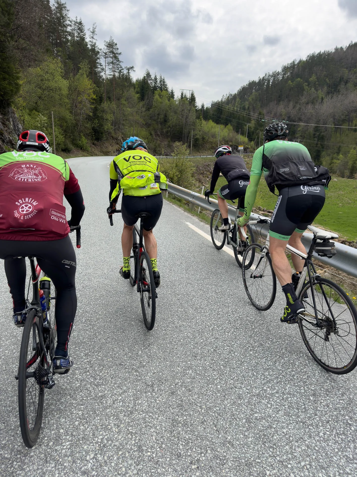 Fire syklister sykler opp en fjellvei omgitt av grønne trær og fjell, med et overskyet himmel.