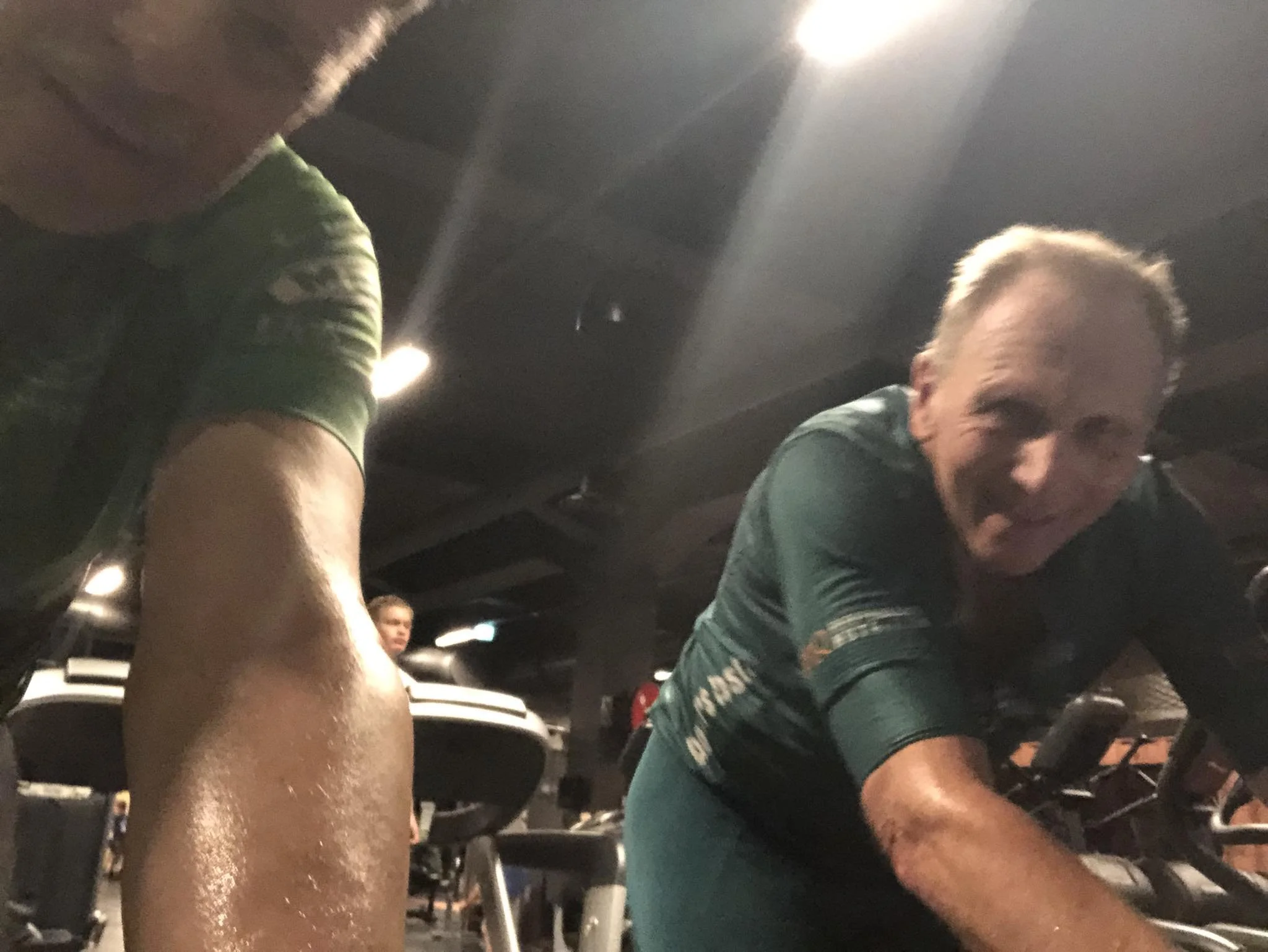 To personer på et treningssenter, en mann foran og en kvinne til venstre. Begge svett, iført treningsklær, ser ut til å trene.