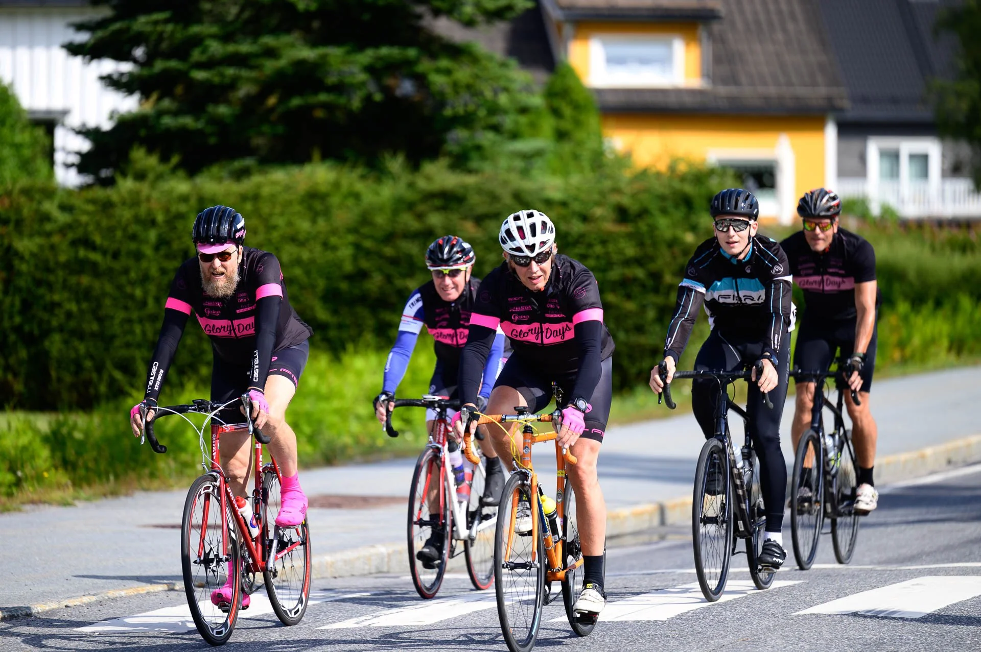 Fem personer sykler på en vei, alle bruker hjelmer og sykler i ulike farger. To av personene har fargerike sykler og klær, alle er i bevegelse, og det er grønne busker og hus i bakgrunnen.