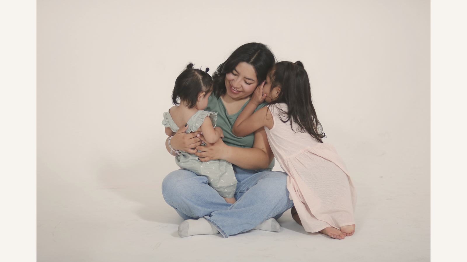 A woman sitting cross-legged on the floor with two young girls, all hugging and whispering happily against a plain white background.
