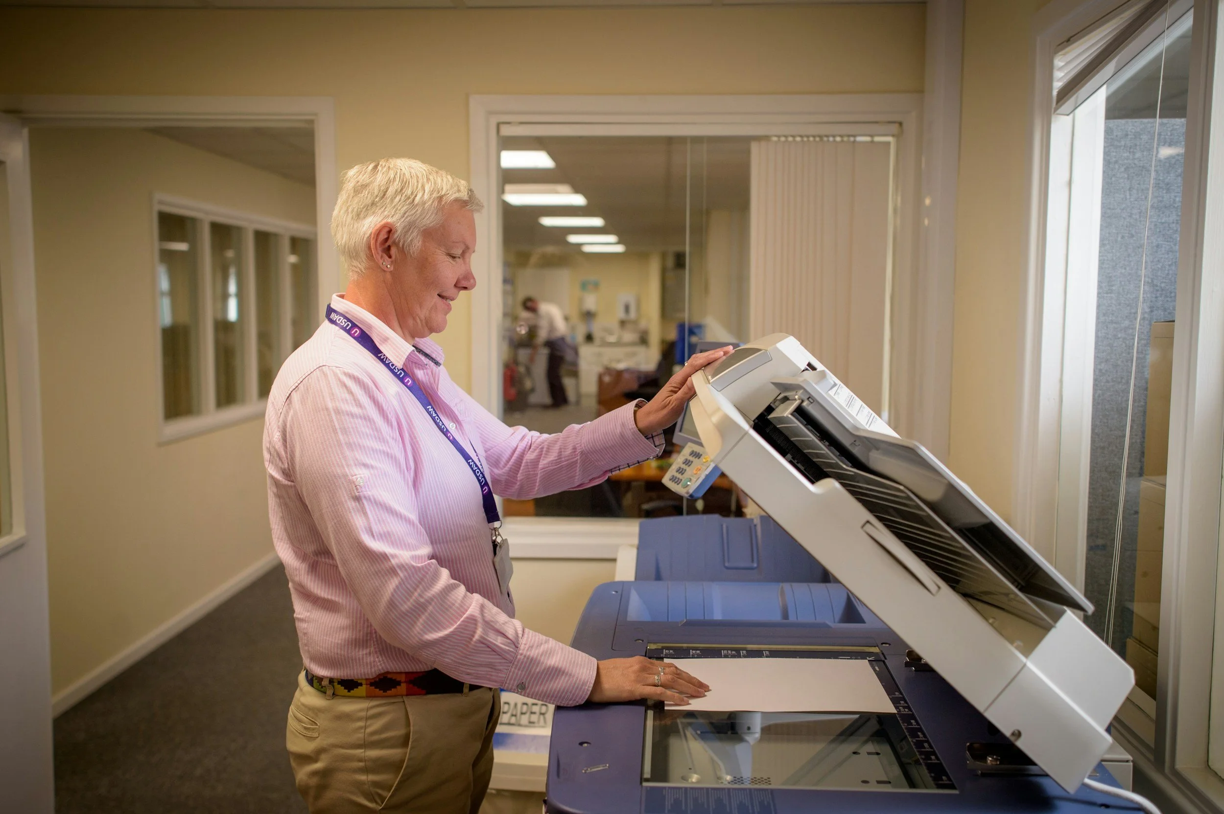 A woman in a pink striped shirt and beige pants using a photocopier or scanner machine in an office environment.