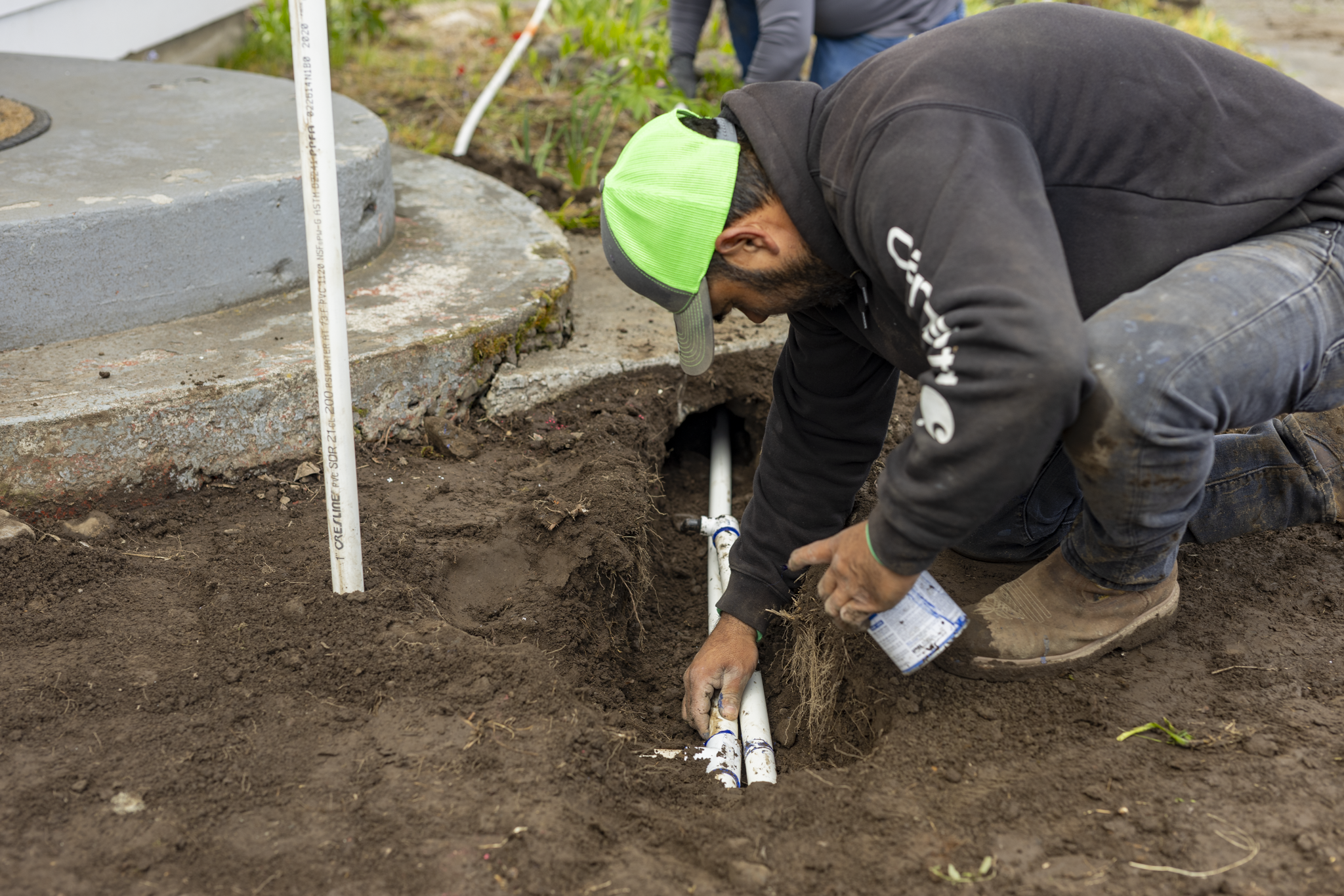 A person wearing a green cap and dark clothing is installing or repairing white pipes in the dirt at a construction site near a concrete structure with plants in the background.