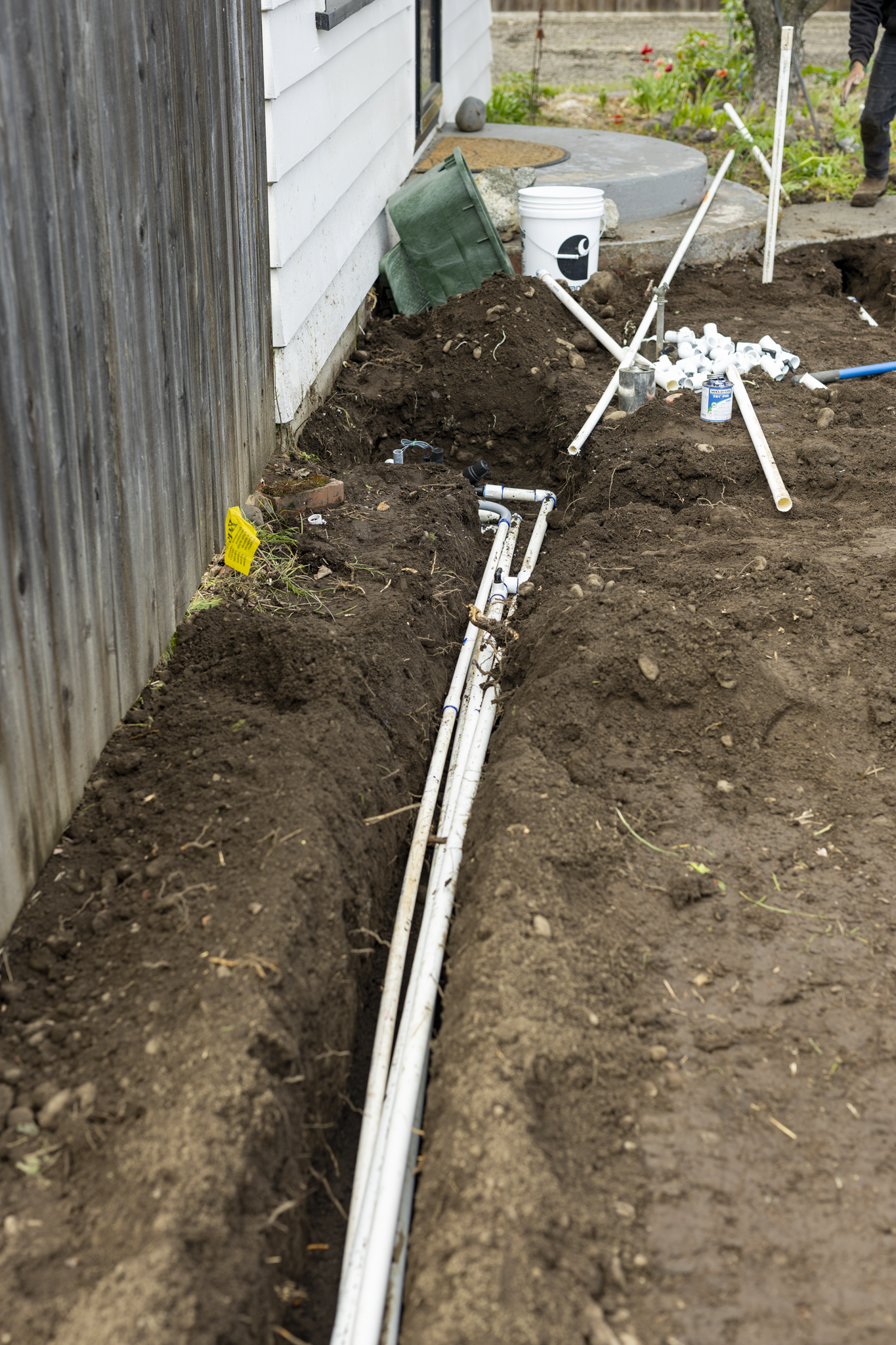 Underground water pipes installation in a trench along the side of a house with white siding, construction tools, and a worker in the background.