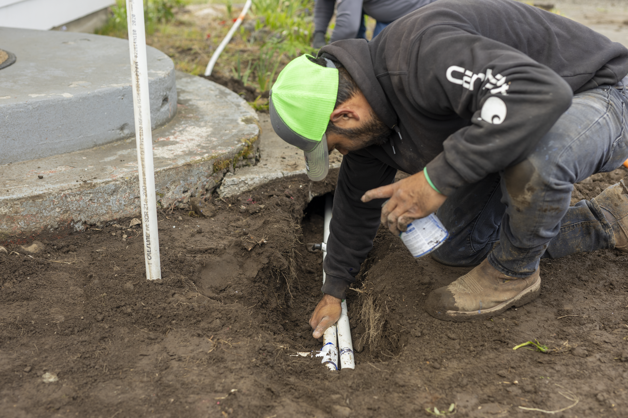 Worker installing underground pipes in a trench, digging along a building foundation, wearing a green cap and dark clothing.