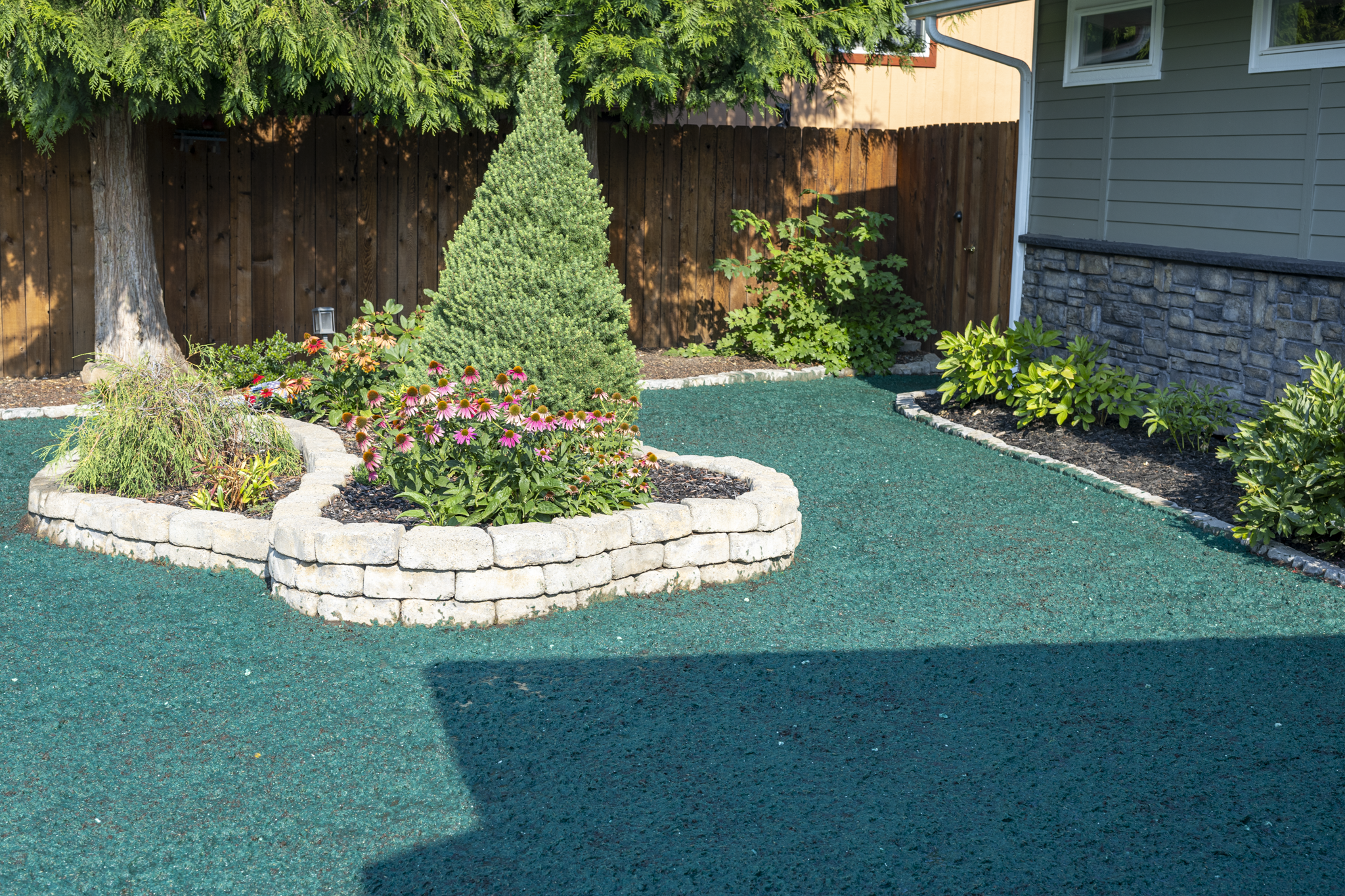 A landscaped yard with a circular flower bed edged with light gray bricks, containing pink and orange coneflowers, with a small cone-shaped evergreen tree in the center, a large tree with green foliage, and various other plants, all bordered by a dar