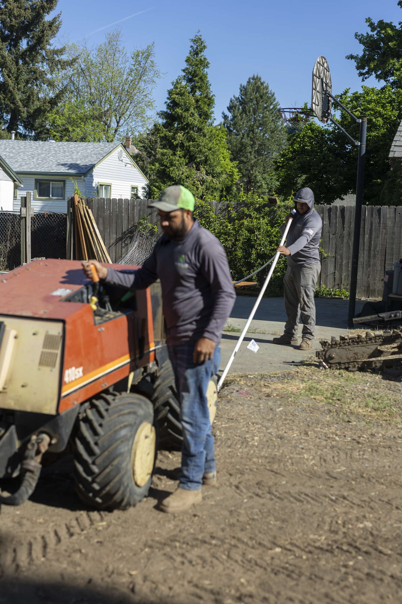 Two workers are engaged in construction or landscaping work in a backyard. One is operating a piece of machinery, while the other holds a long white pipe. There is a basketball hoop mounted on a pole, a wooden fence, and trees in the background under