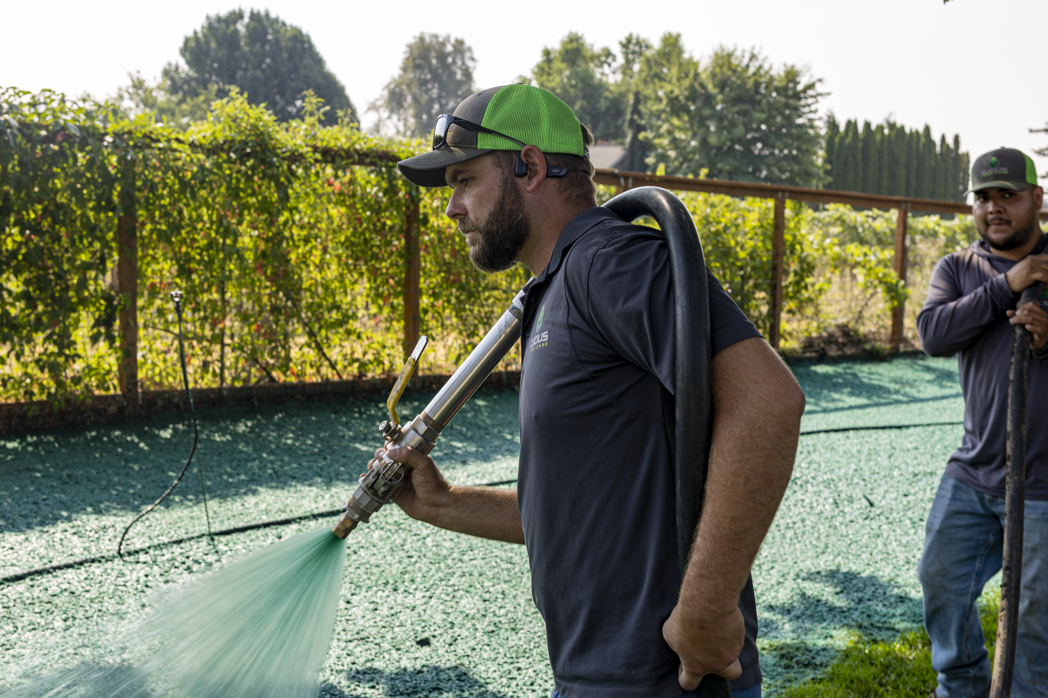 A man wearing a green and black baseball cap and black shirt is holding a tool and sprinkling a substance onto a green surface outdoors. Another man in a gray cap and dark hoodie is standing to the right, partially visible. There is green foliage and