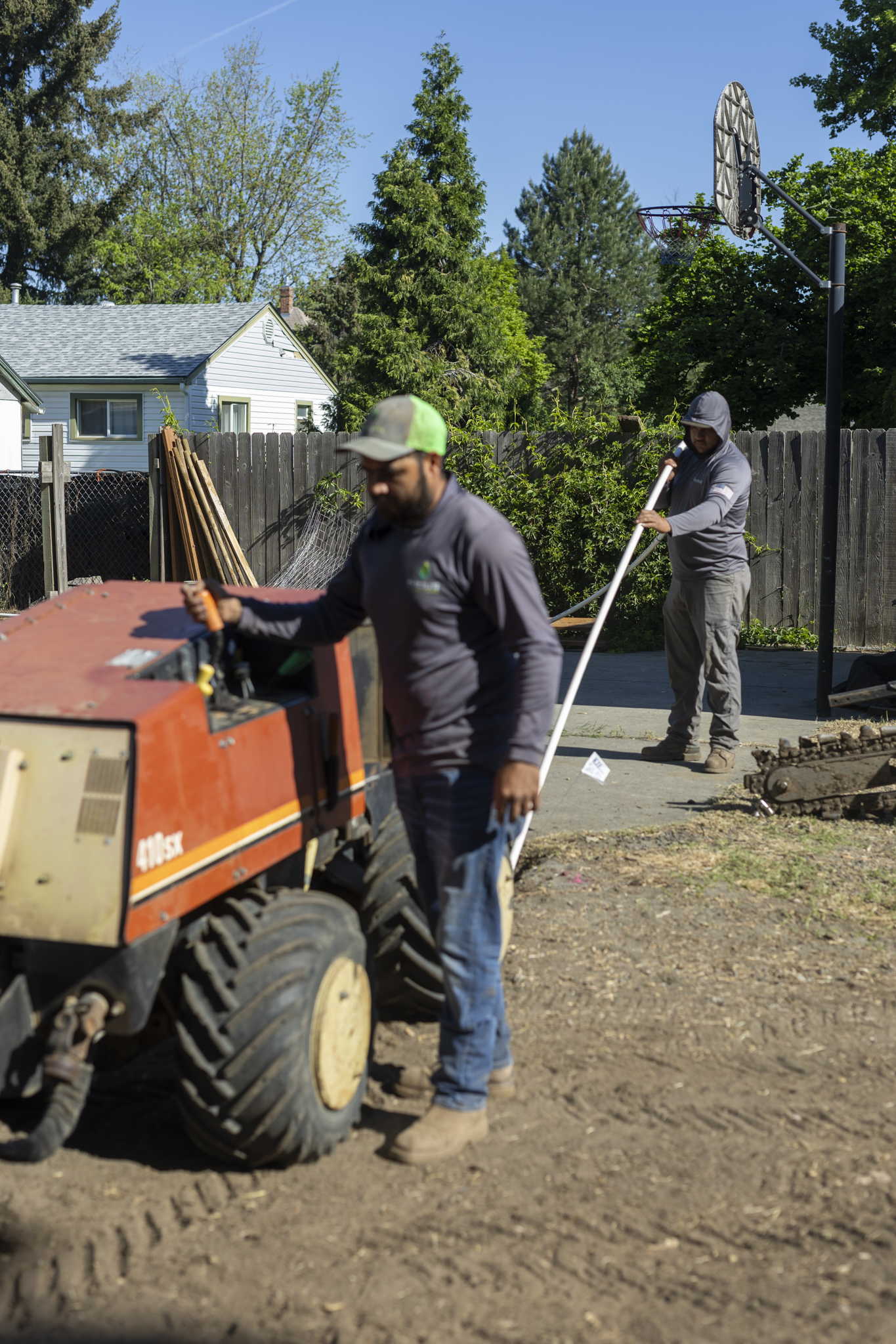 Two workers operating heavy equipment in a yard, with trees and a house in the background, under a clear blue sky.