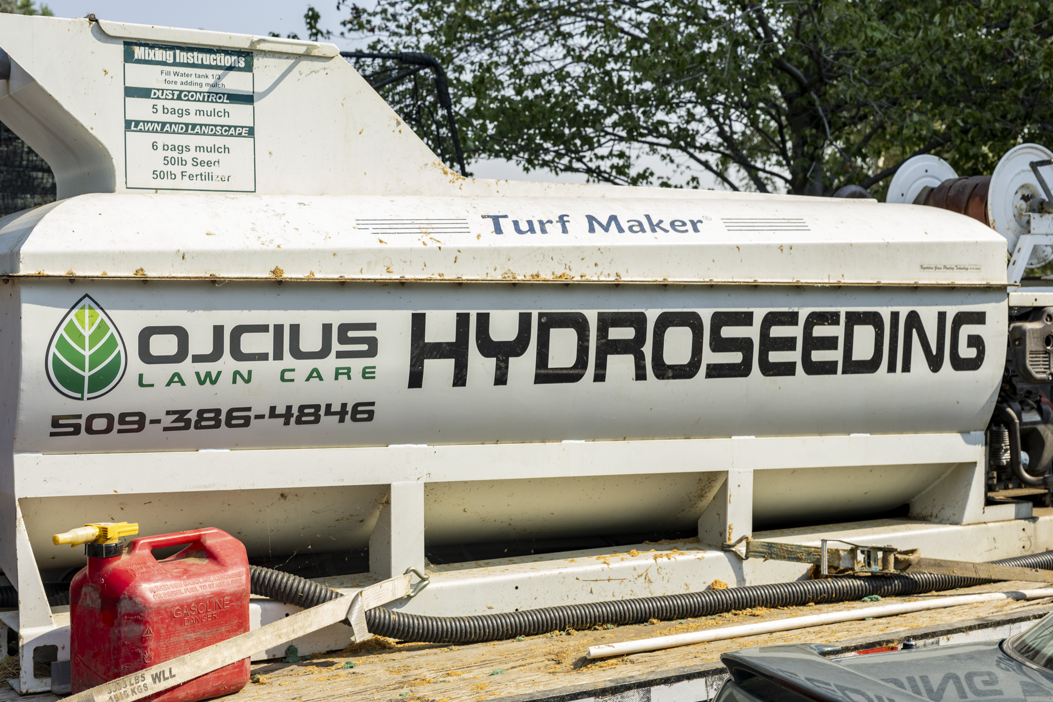 A large white hydroseeding machine with green and black text, reading 'OJCUS Hydro Seeding Lawn Care' and a phone number, with a red gas can in front and green trees in the background.