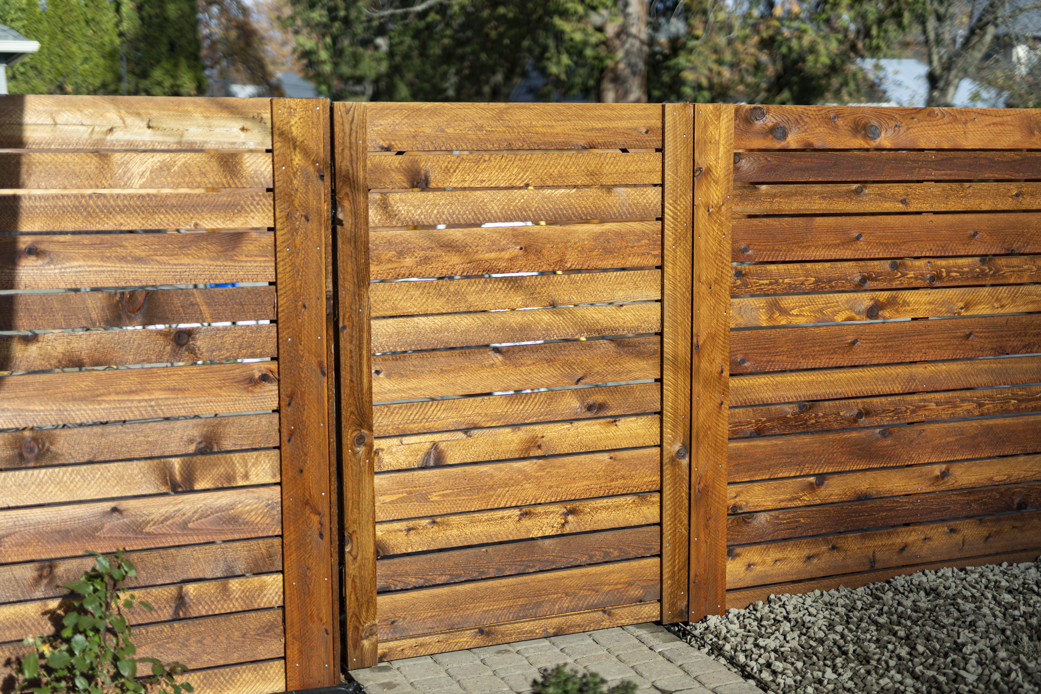 Wooden privacy fence with horizontal planks and vertical support beams, with a driveway and trees in the background.