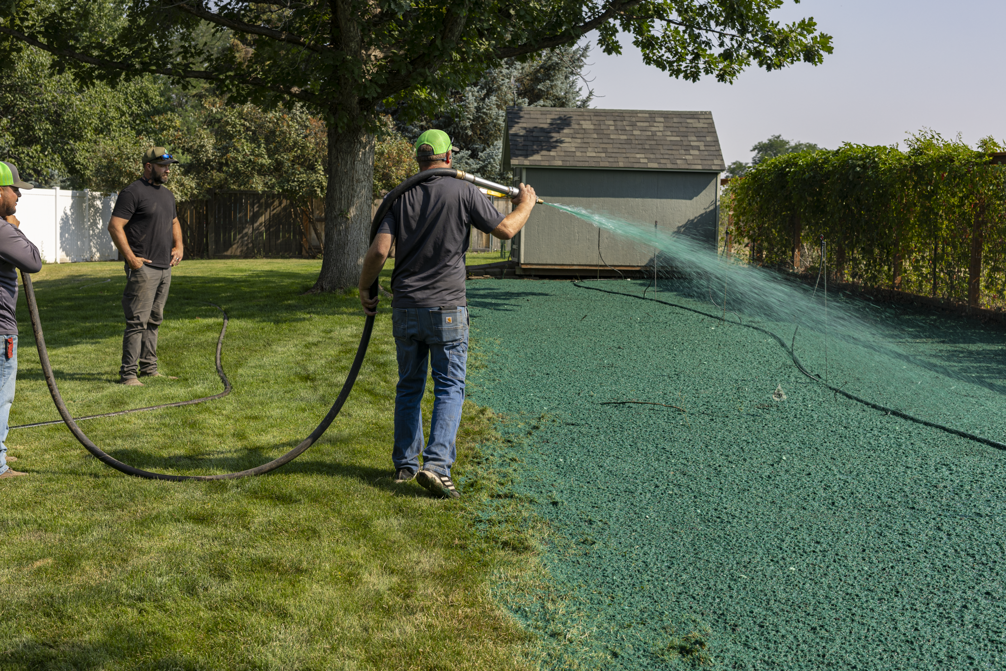 Three men watering a backyard with a garden hose while standing on green grass, with a shed, trees, and a wooden fence in the background.