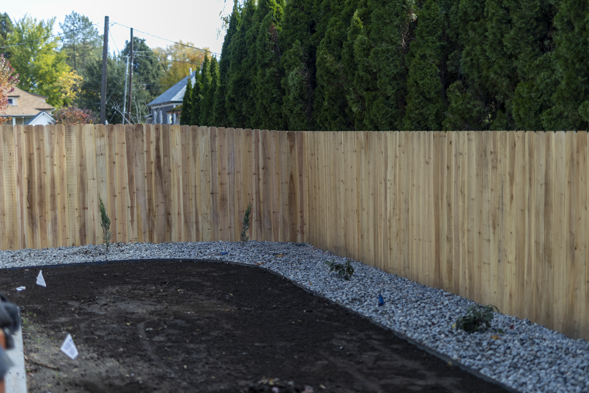 A newly built wooden fence surrounds a garden bed with dark soil and small plants. The ground is covered with gravel, and there are small flags marking the planting areas. Trees and houses are visible in the background under a clear sky.