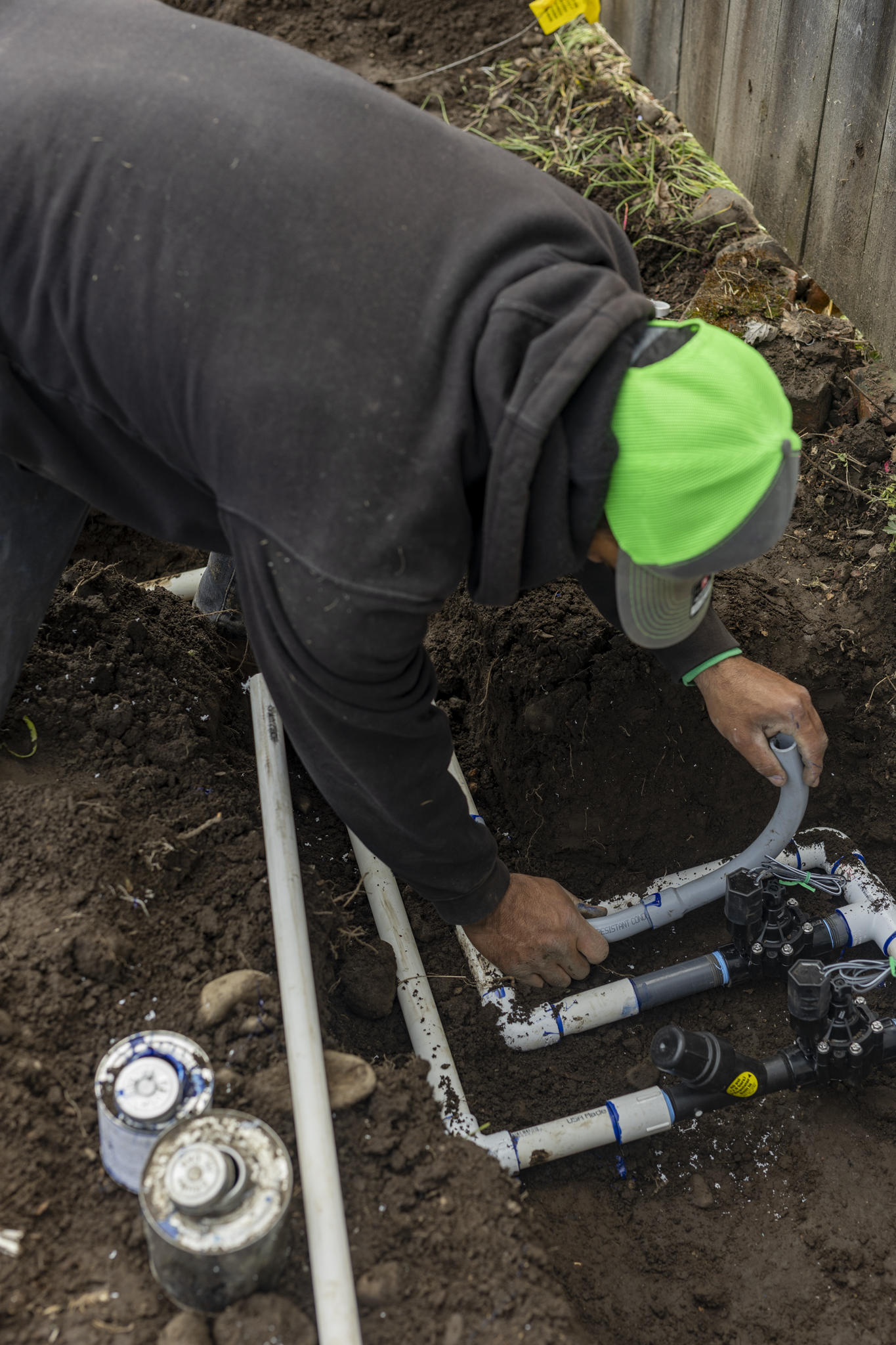 A person wearing a black jacket, green hat, and gray cap is installing or repairing underground plumbing pipes in a garden or yard area, with soil and a wooden fence visible.