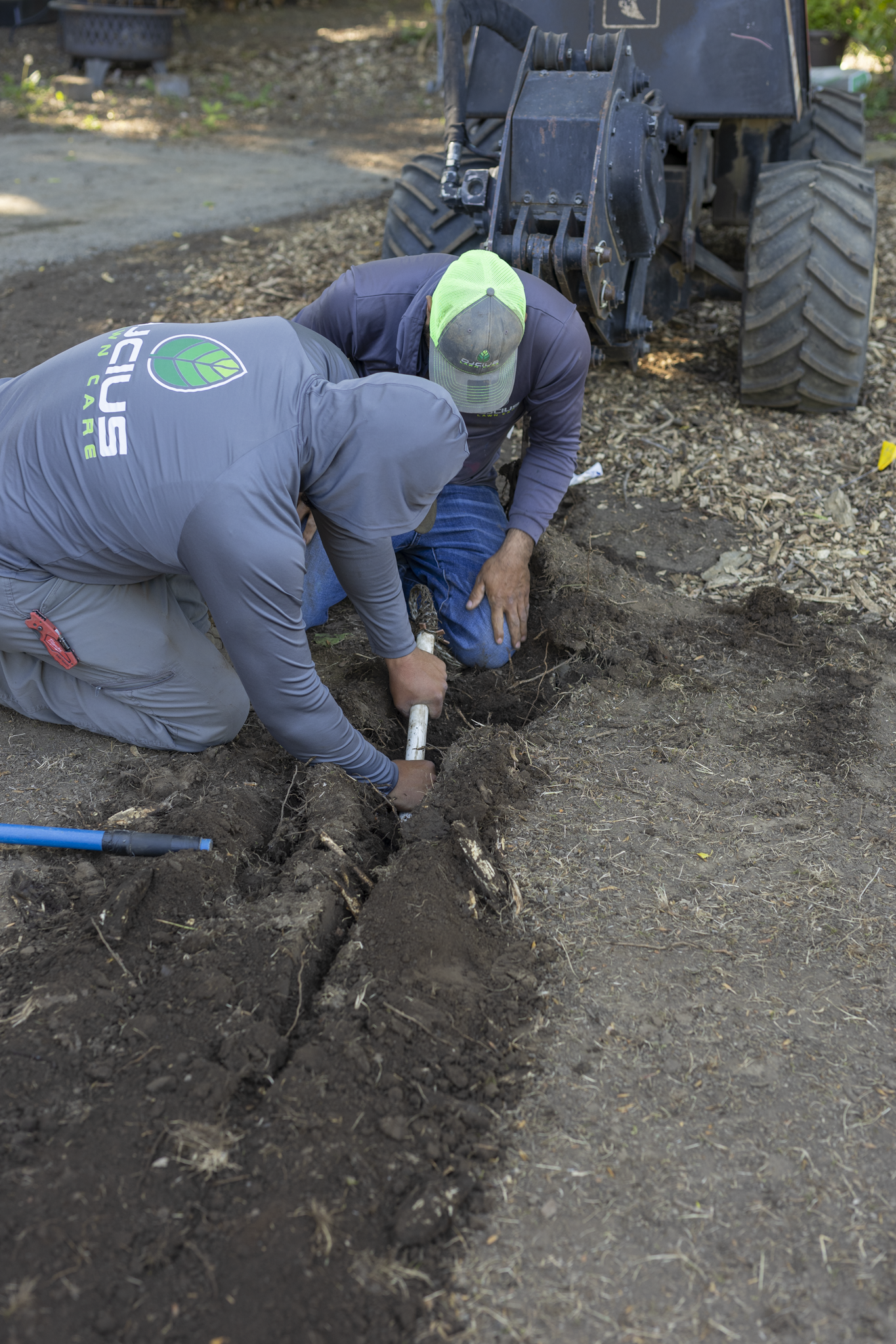 Two individuals working together to dig a hole in the ground using a shovel near machinery, possibly in an outdoor work site.