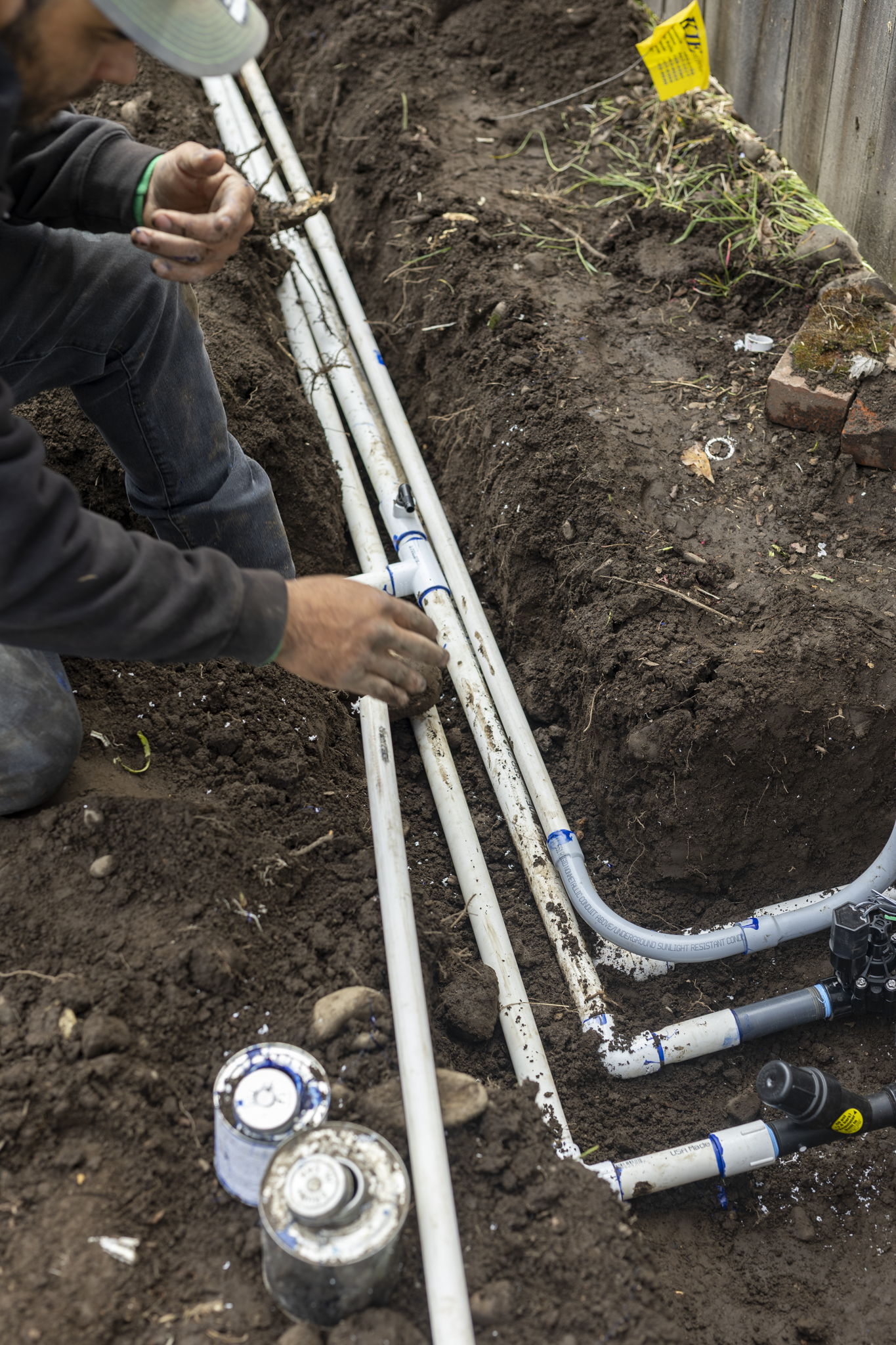 A person working on installing or repairing white PVC plumbing pipes in a trench dug in the dirt. The person is wearing a gray baseball cap, a black hoodie, and blue jeans. There are cans of paint or sealant cans placed in the foreground, and a yello