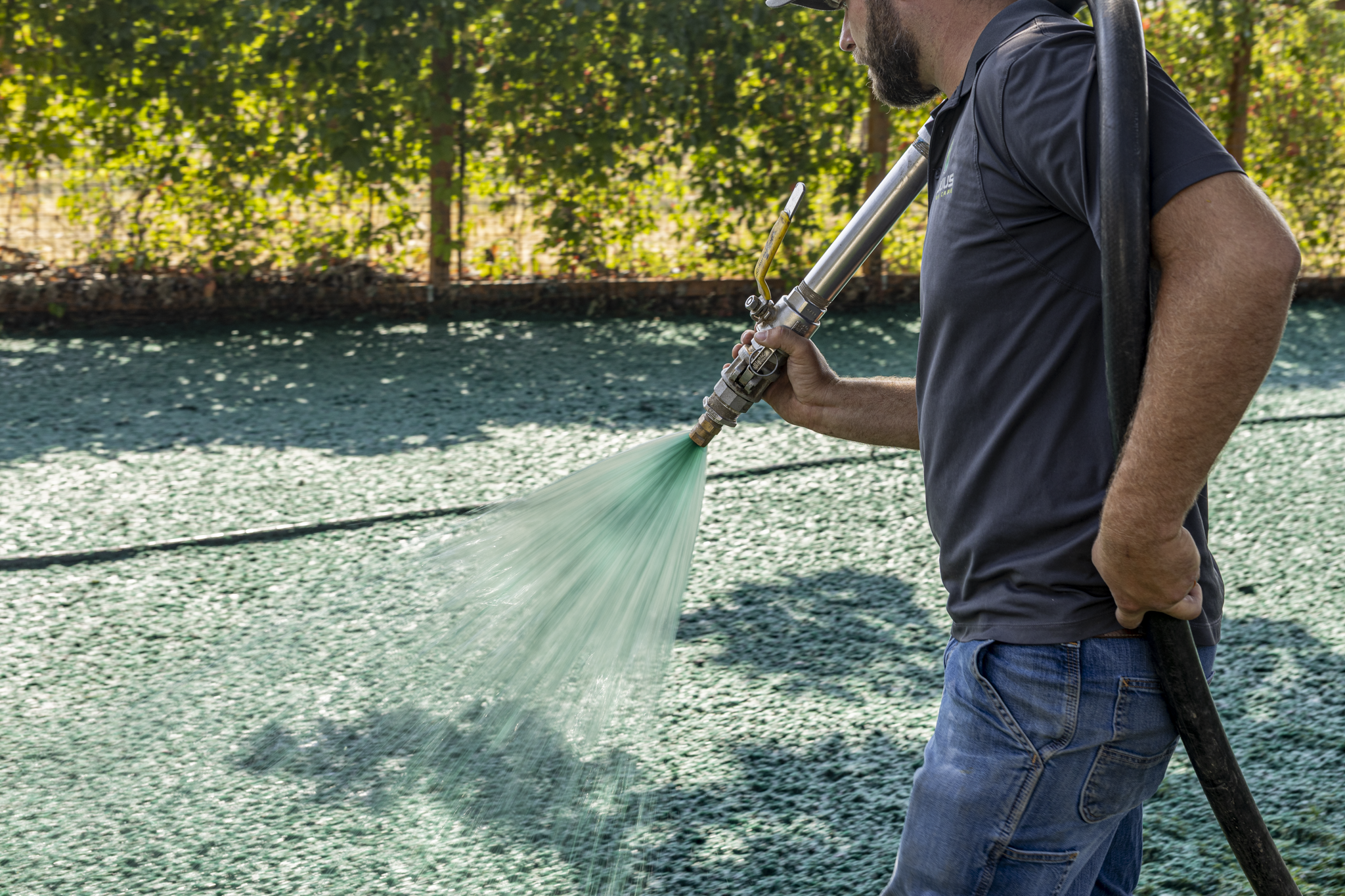A man with a beard wearing a dark polo shirt and jeans holding a spray nozzle, watering a large outdoor area with green gravel during daytime.