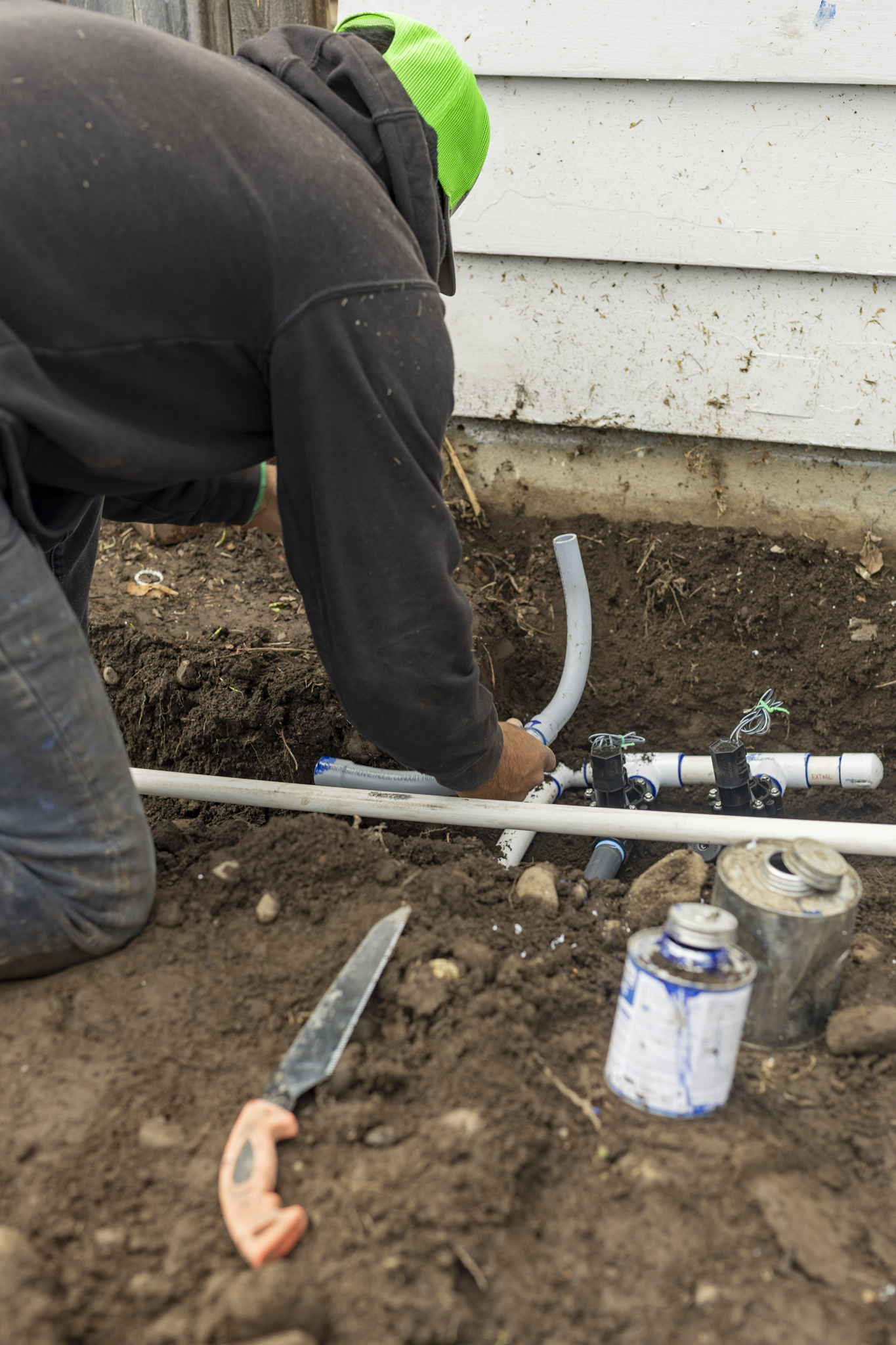 A person wearing a green hat and black hoodie installing plumbing pipes in a dirt-covered ground outside a house with white siding.