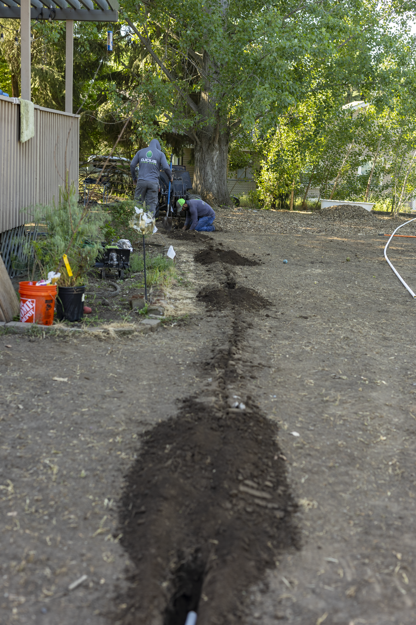Two workers are digging a trench in a backyard with a large tree and green foliage in the background. One worker is kneeling while the other is standing, both focused on the dirt. Garden supplies and tools are visible around them.