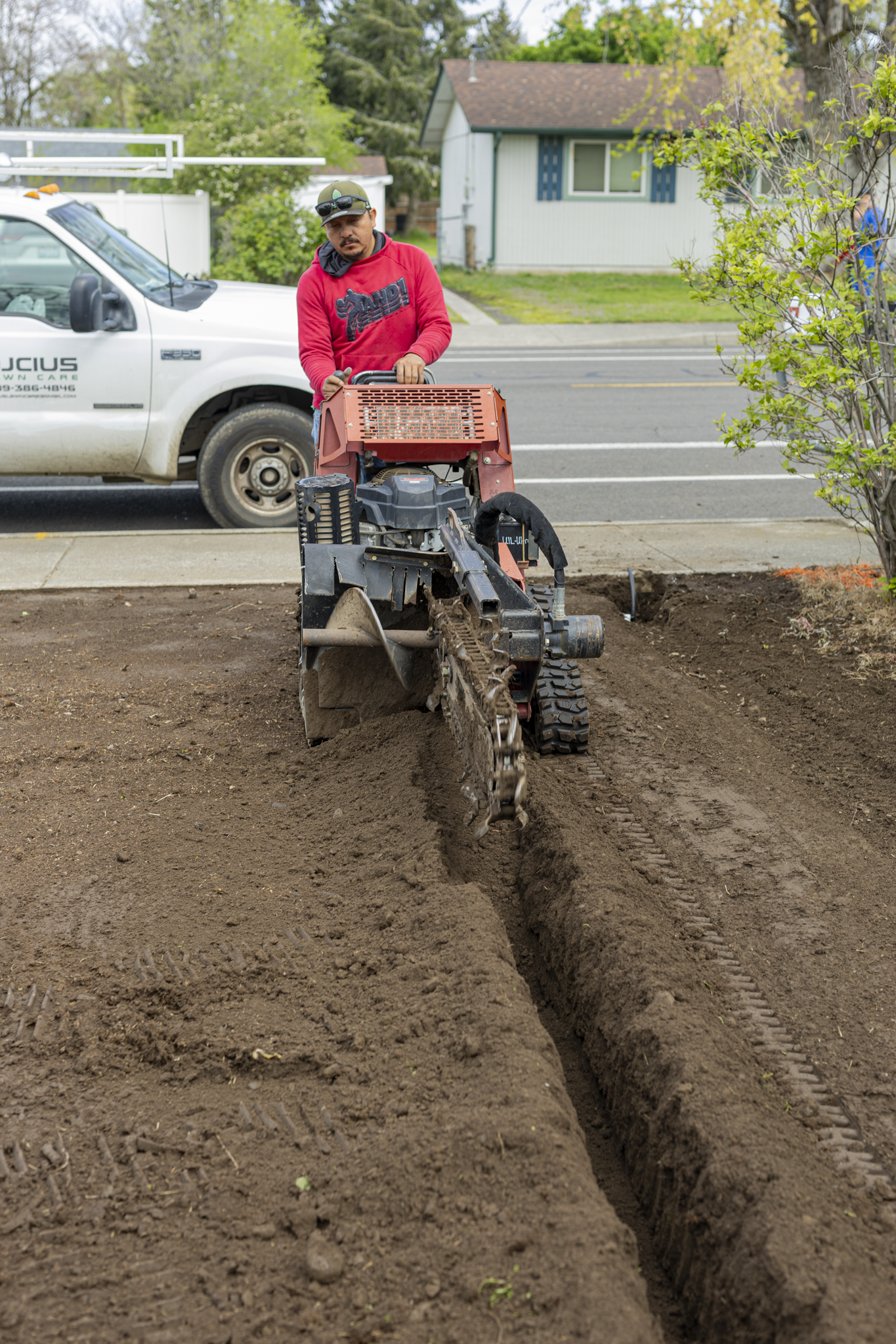 Man operating a trench compactor in a residential yard, with a white truck and house in the background.