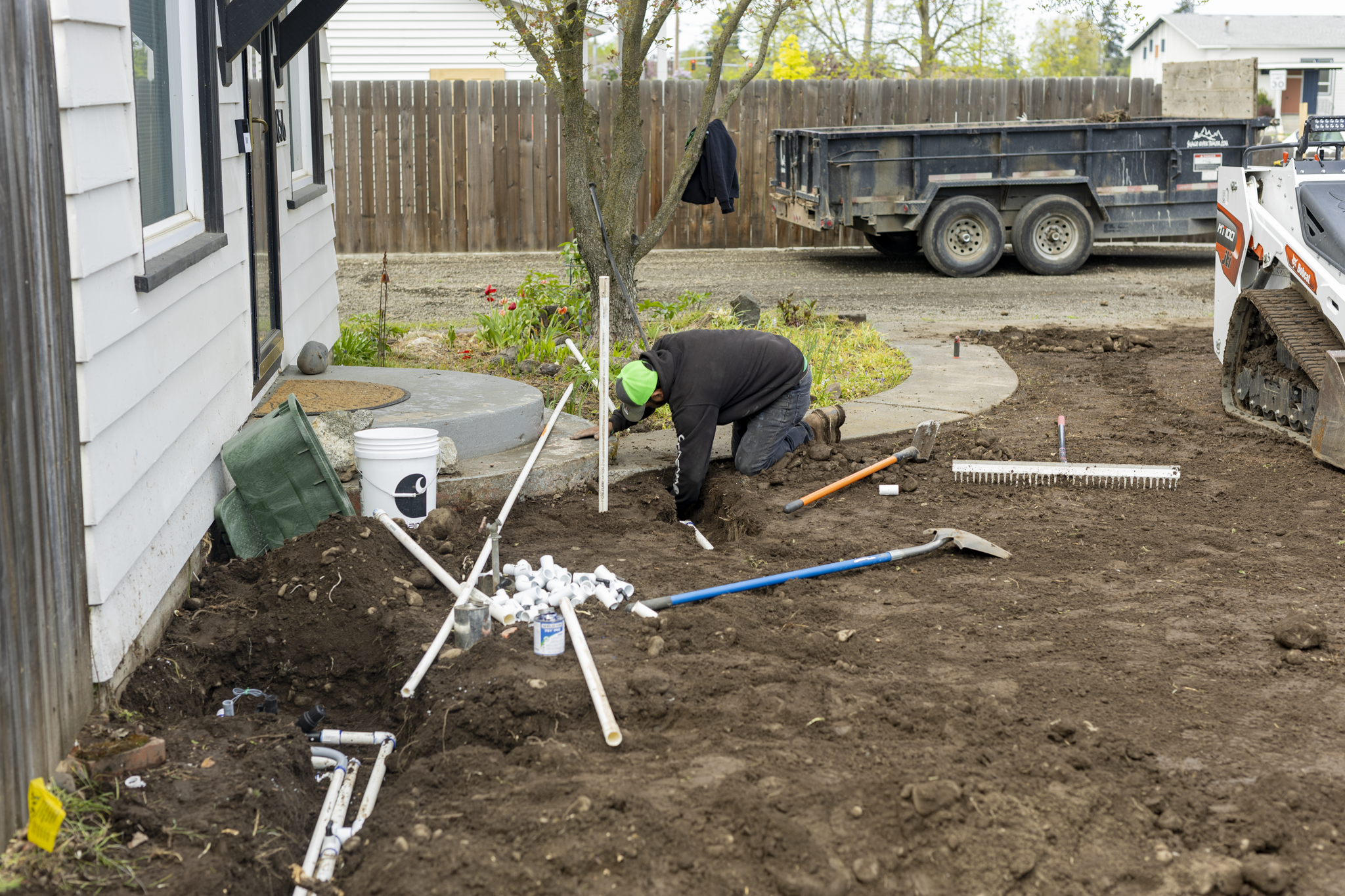 Construction workers installing underground piping in a backyard. One worker is kneeling digging, while another is working near a tree. A dump truck and a mini excavator are present, with various pipes and tools scattered around.