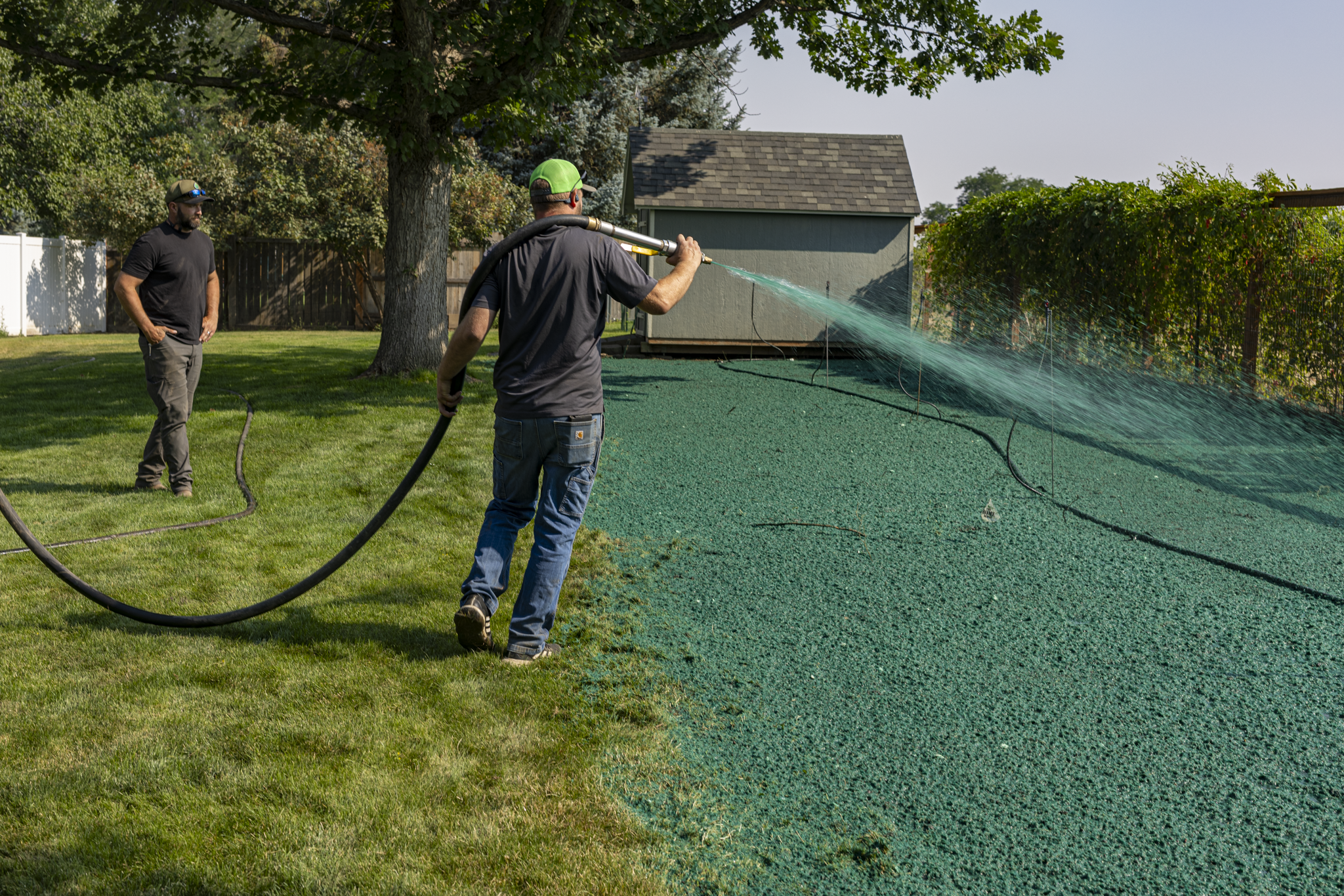 Two men in a backyard are watering a green turf with a garden hose. One man is standing, the other is spraying water on the ground. Trees and a shed are visible in the background.