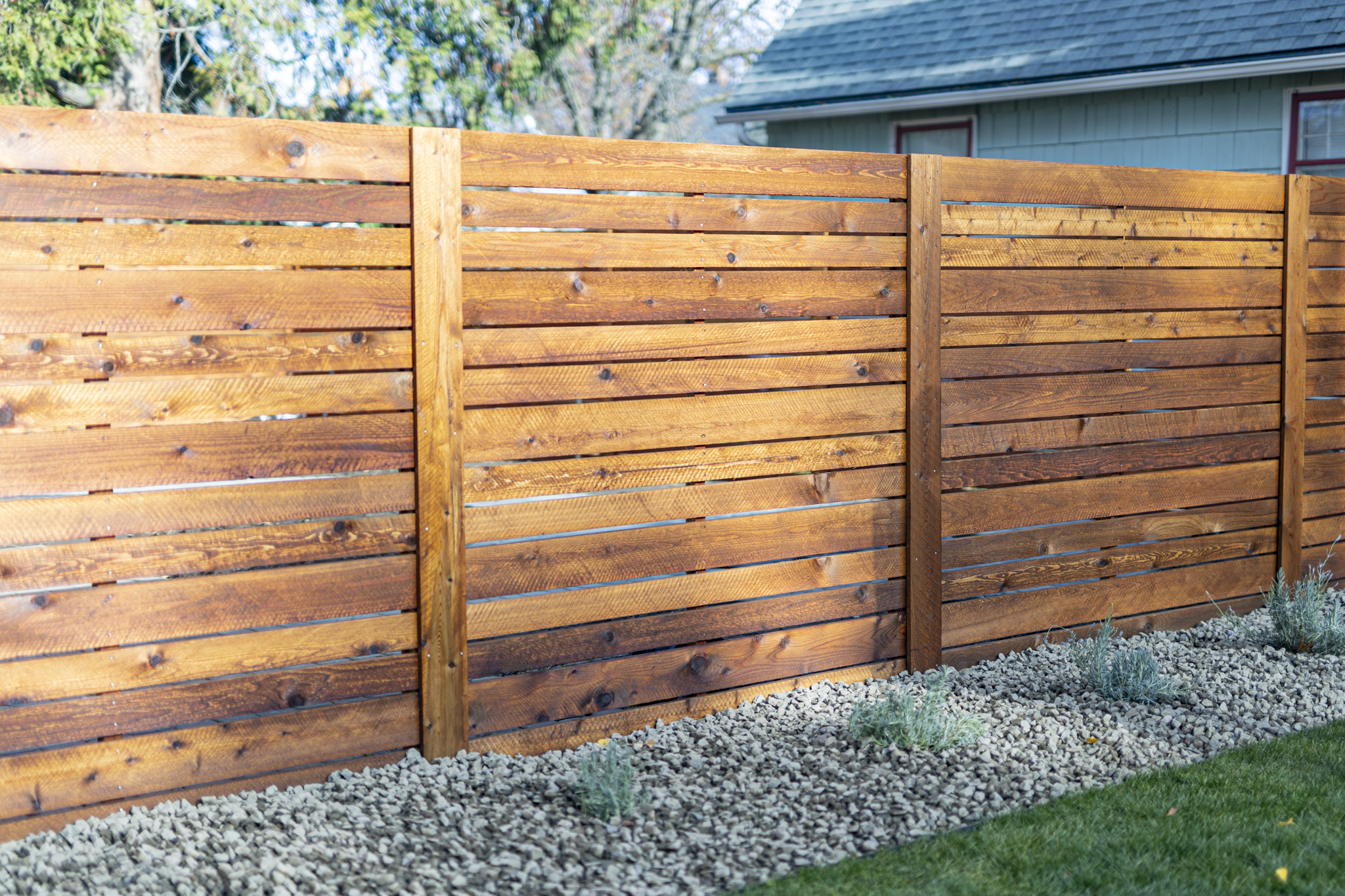 A wooden fence with horizontal planks and vertical support posts, with a house and trees in the background and a bed of rocks and small plants at the base.