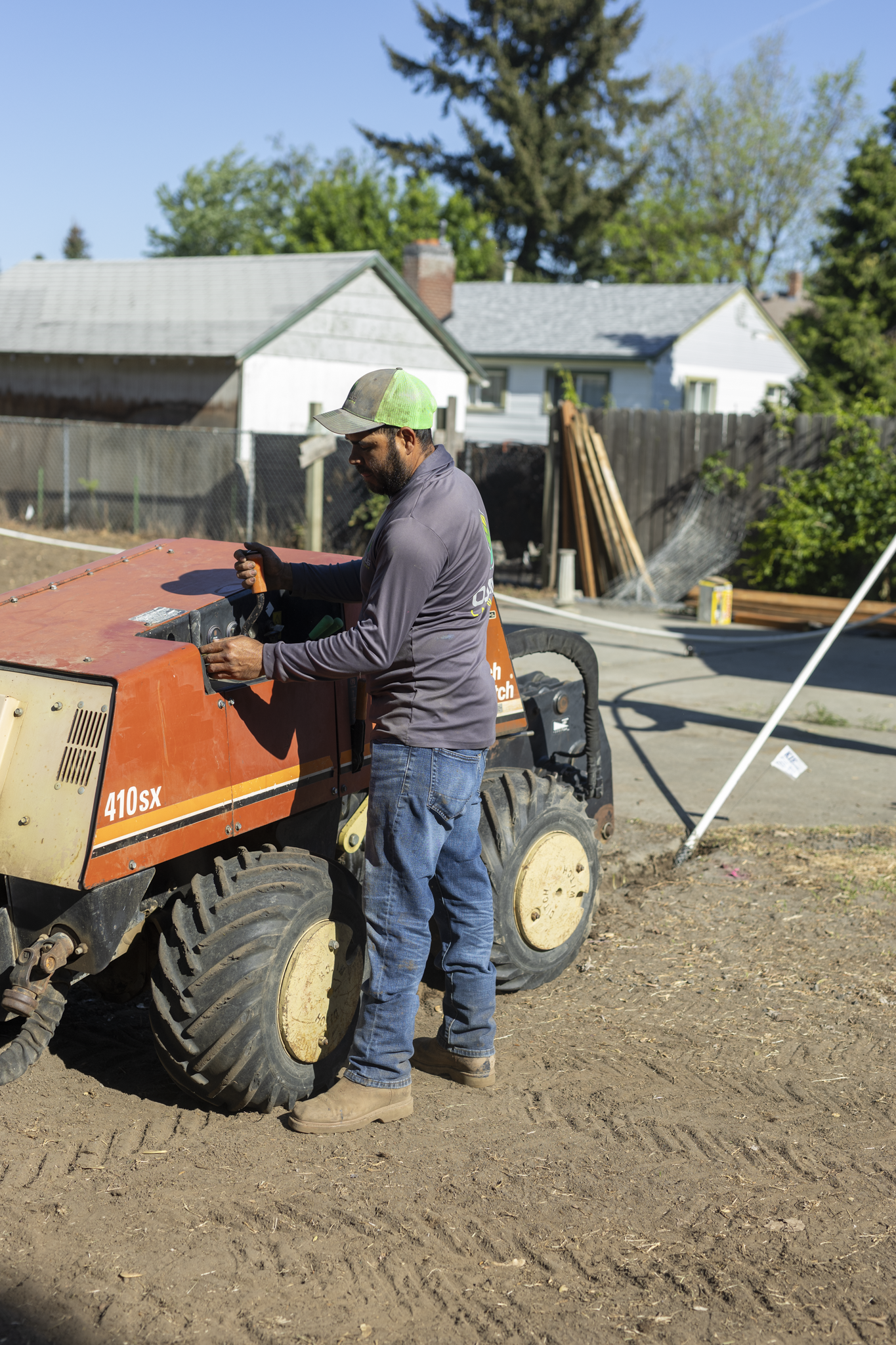 A man wearing a green and gray trucker hat, gray long-sleeve shirt, jeans, and boots working on a small orange construction vehicle labeled '410SX' outside in a fenced backyard with trees and a white house in the background.