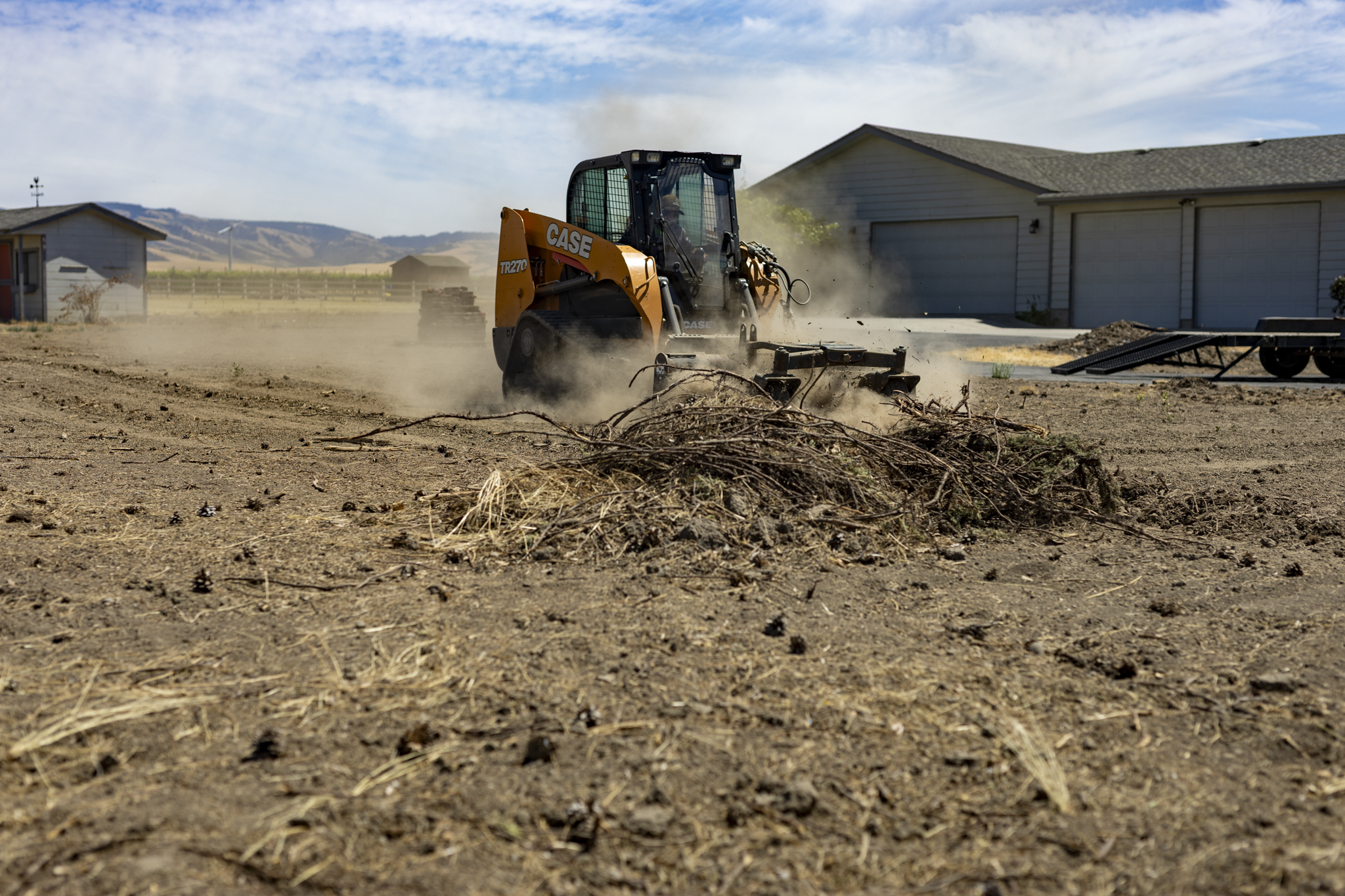 A small orange and black CASE ride-on turf grader smoothing dirt ground, with dust clouds behind it, in a suburban backyard with houses and mountains in the background.