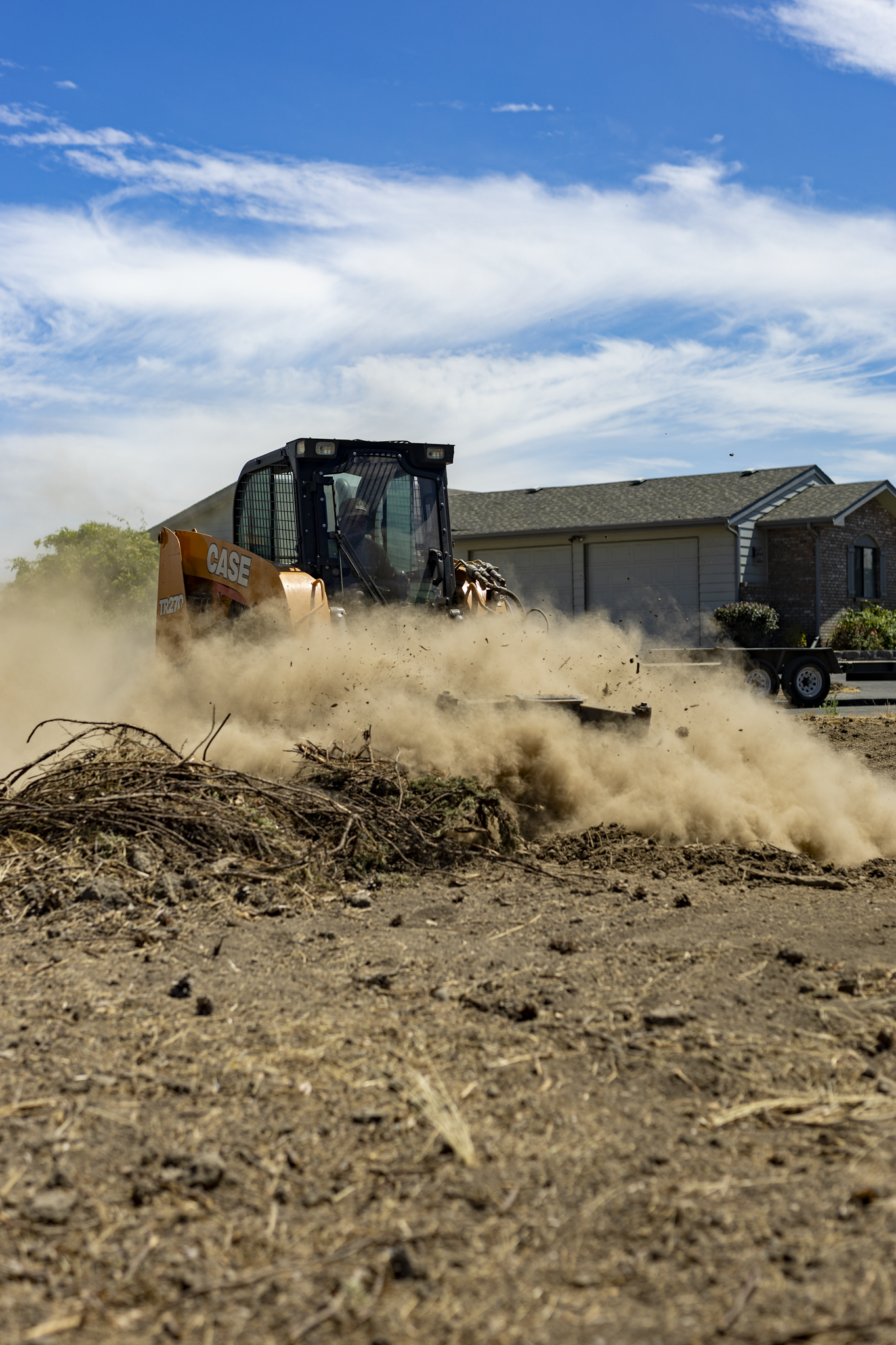 A construction vehicle bulldozer spreading dirt and debris on a dirt lot. There are houses in the background and a cloudy blue sky above.